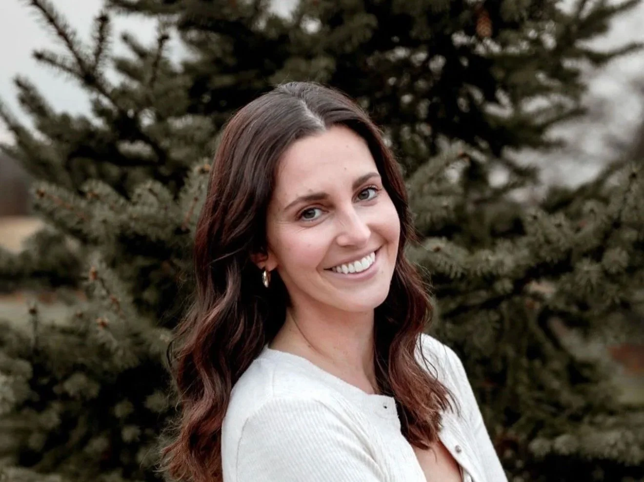 A woman with wavy brown hair and blue eyes, smiling, standing outdoors in front of a pine tree, wearing a light-colored top and earrings.