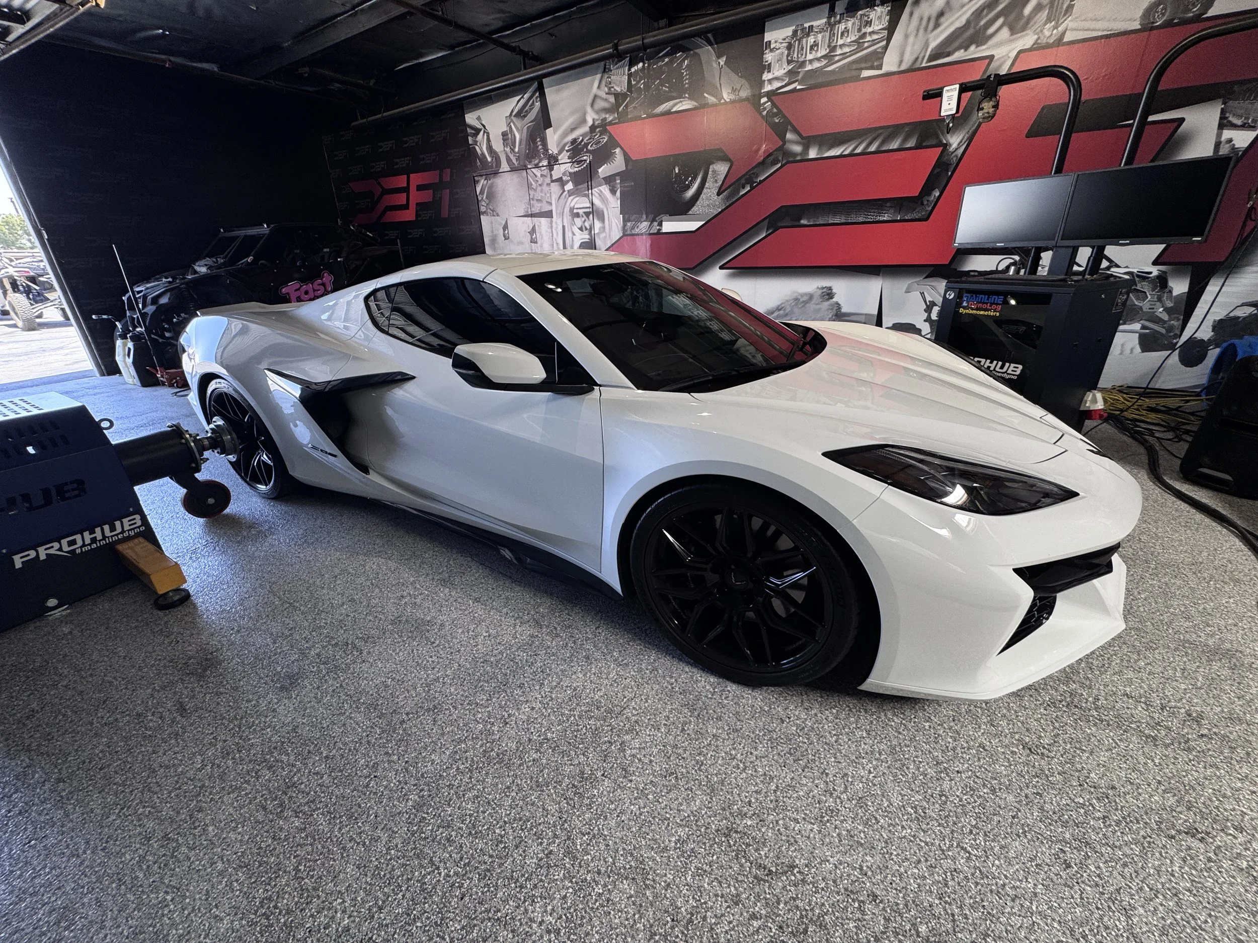 A white sports car is positioned inside a racing simulation setup with two monitors on a stand and a black racing seat in the background, all inside a room with black and red decor.
