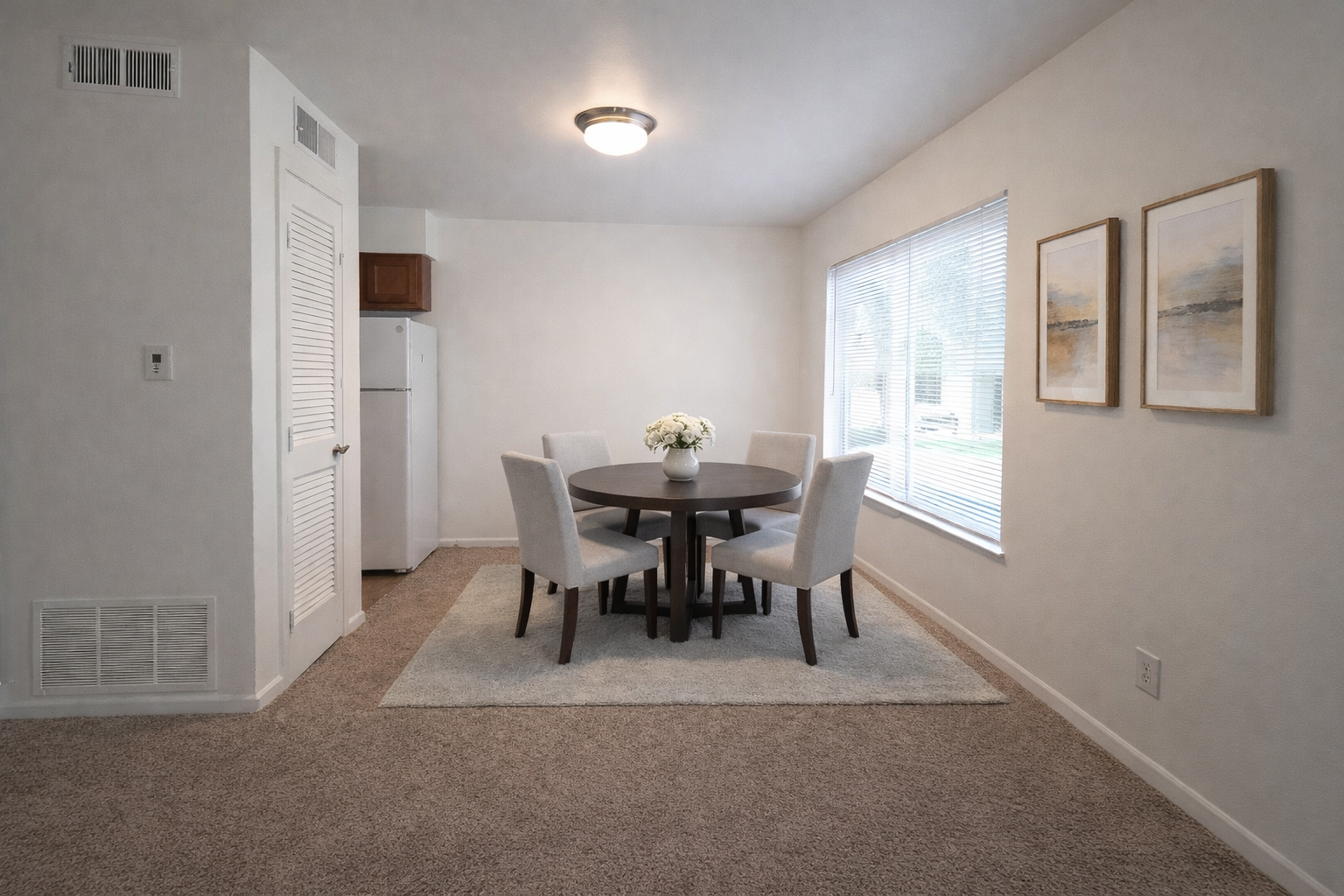 Dining area with a round table and four upholstered chairs, a vase of white flowers on the table, large window with blinds, framed artwork on the wall, and a small rug beneath the table, with a kitchen refrigerator in the background.