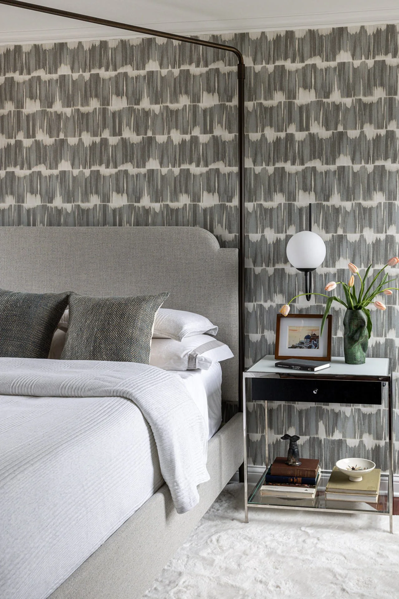 canopy bed with black and white wallpaper and a silver nighstand filled with books and florals.