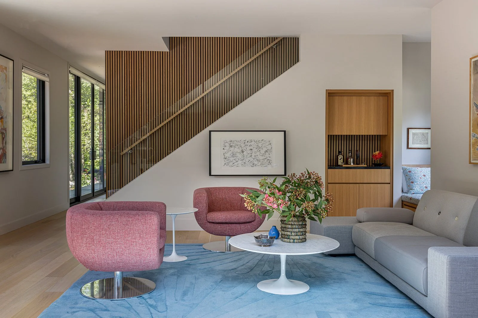 Modern living room with wood slat staircase wall, pink lounge chairs, blue rug, and natural light from floor-to-ceiling windows