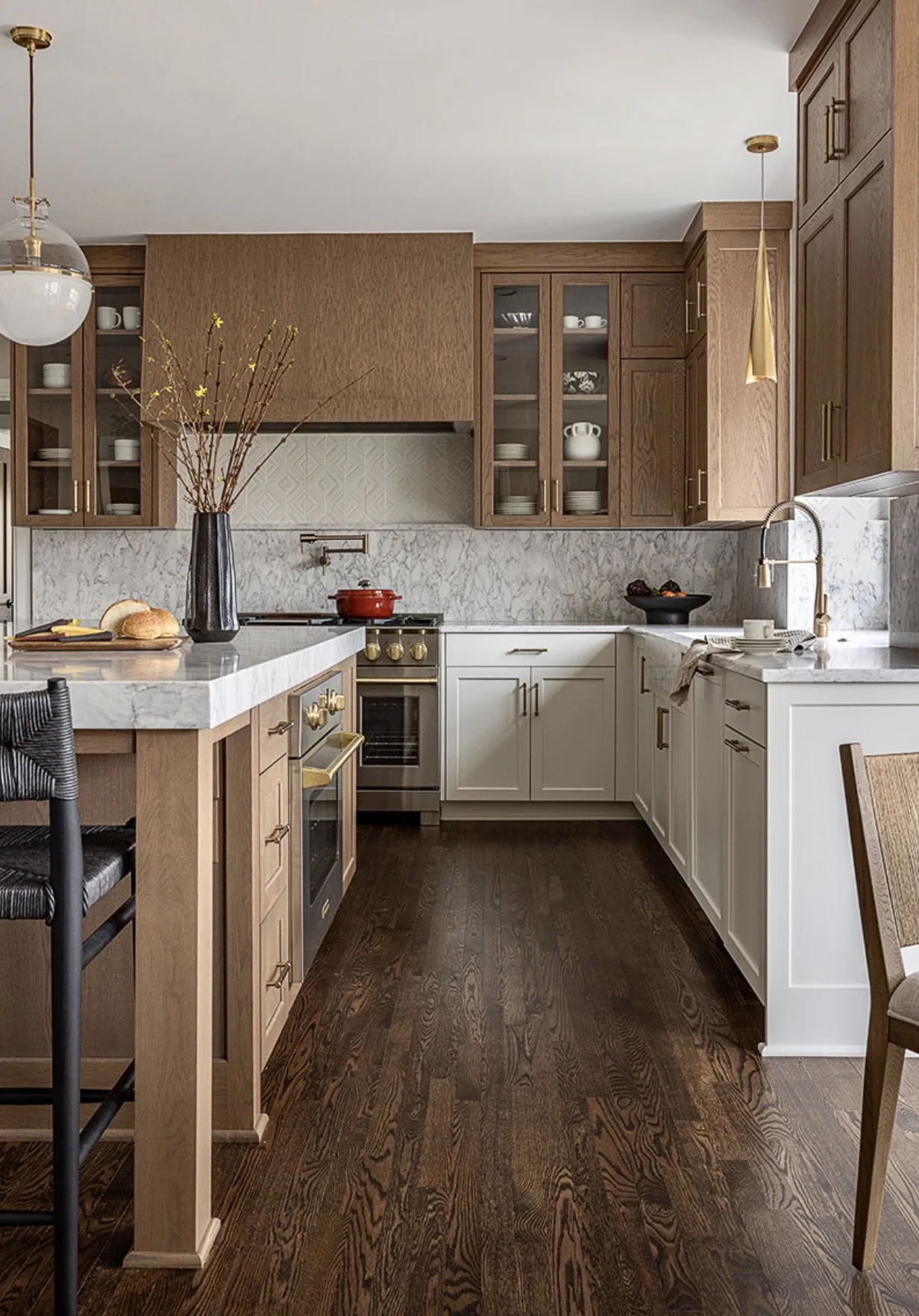 Kitchen with white and wooden cabinets, marble counters, dark wood floor, a vase with branches on the countertop, a red pot on the stove, a black bowl with fruit, a sink with a white cup, and pendant lights.