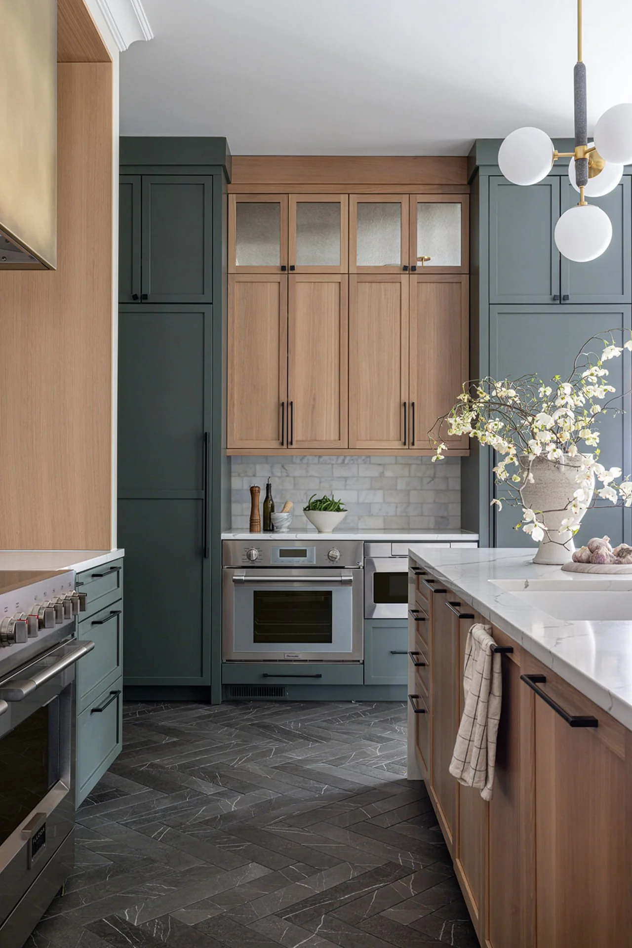 Modern kitchen with green cabinetry, light wood cabinets, marble countertops, and herringbone dark tile floor