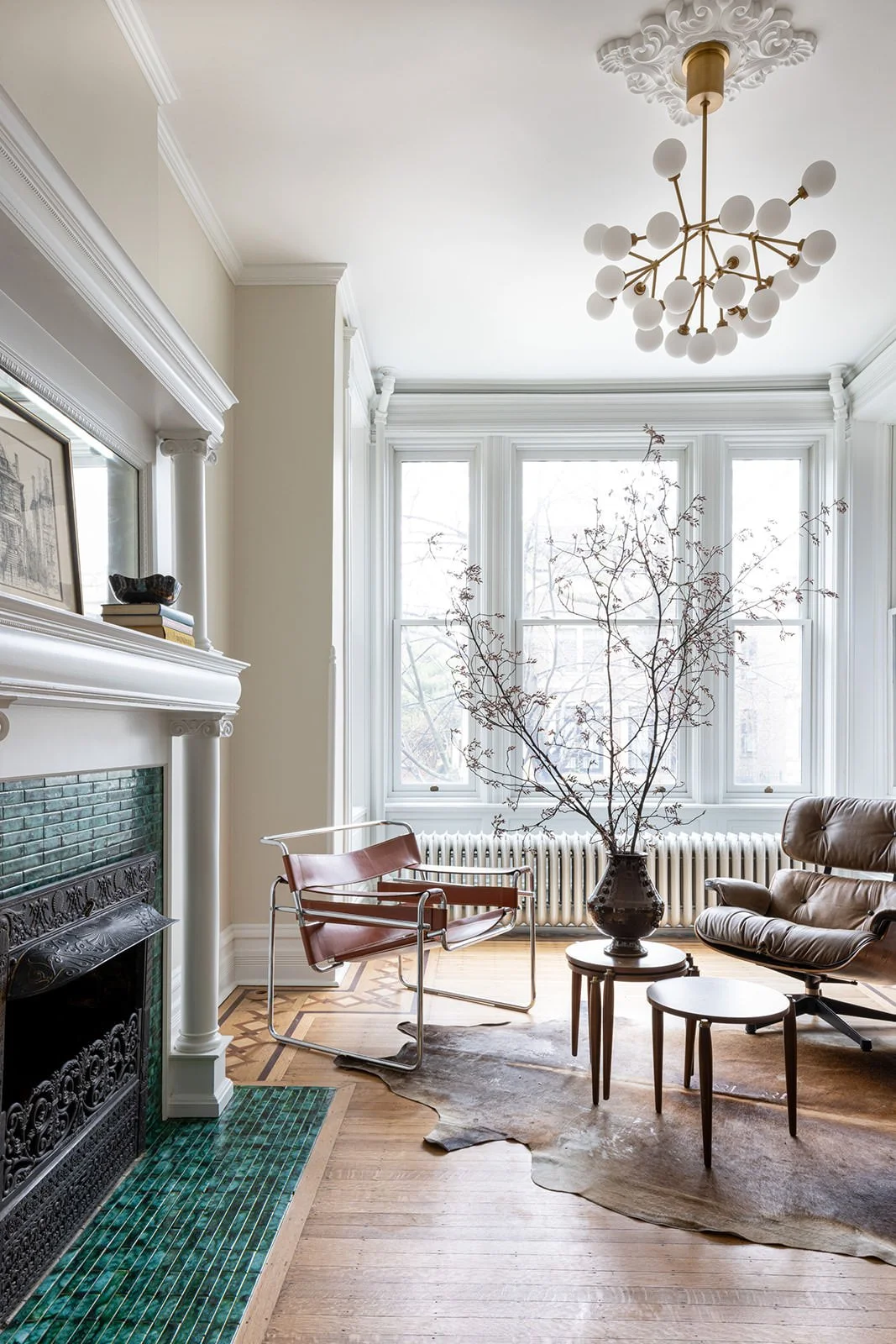Living room with large window, white walls, wood flooring, a black fireplace with green tiles, a brown leather lounge chair, a vintage chair, a round wooden side table, a tall vase with bare branches, and a modern chandelier.
