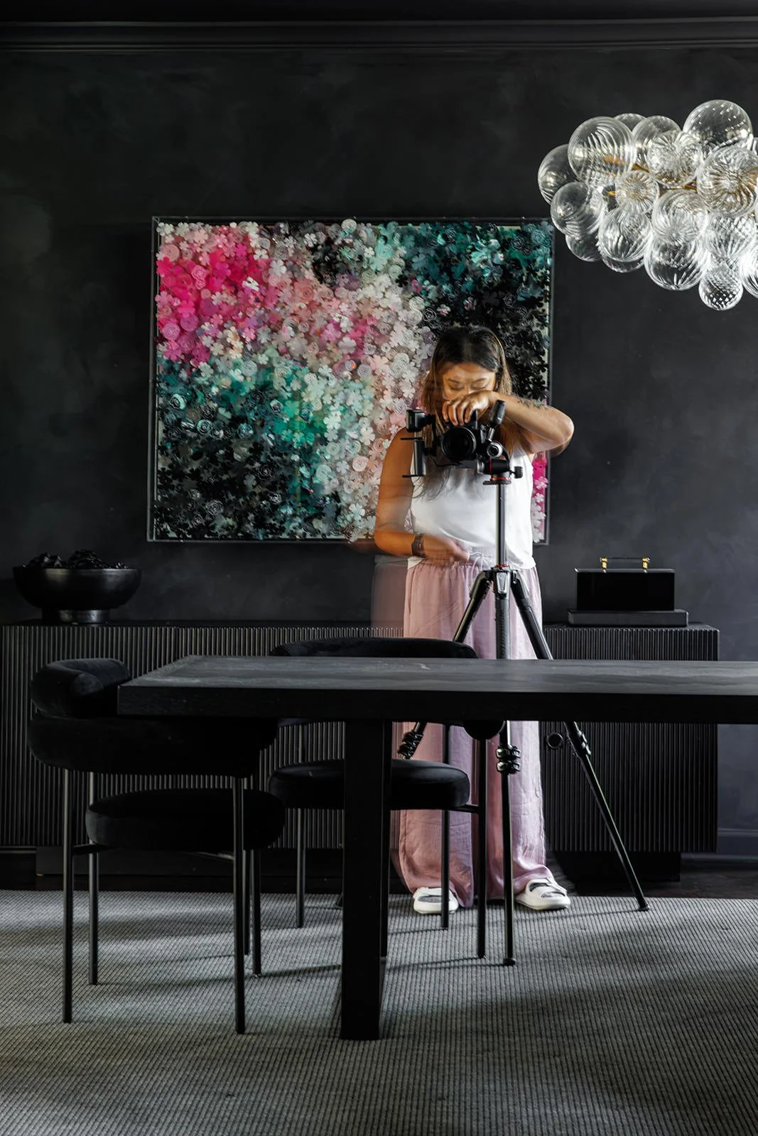 Chicago based interior design photographer taking a photo with a camera on a tripod in a modern room with dark walls, a large colorful floral artwork on the wall, a glass chandelier, a black bowl of fruit, and black furniture.
