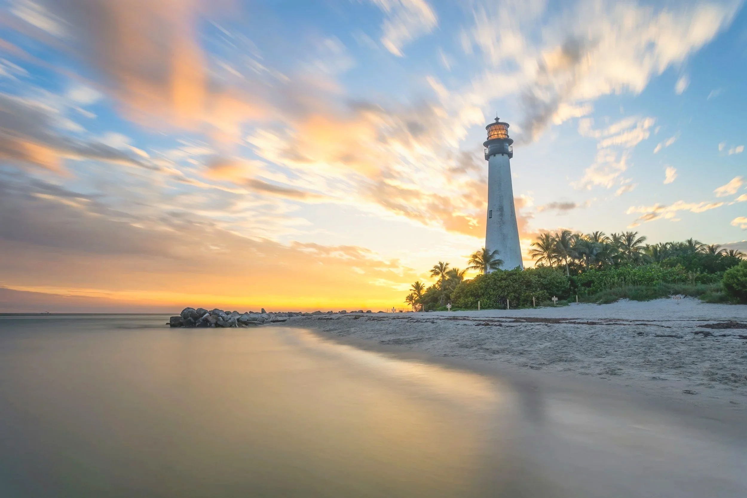 Sunset over the ocean with a lighthouse on a small island, surrounded by palm trees, and a rocky jetty extending into the water.