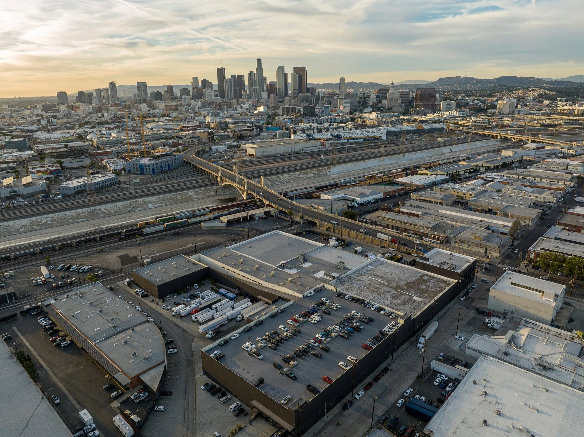 Aerial view of Los Angeles cityscape with highways, train tracks, industrial buildings, and Downtown Los Angeles skyline in the background during sunset.