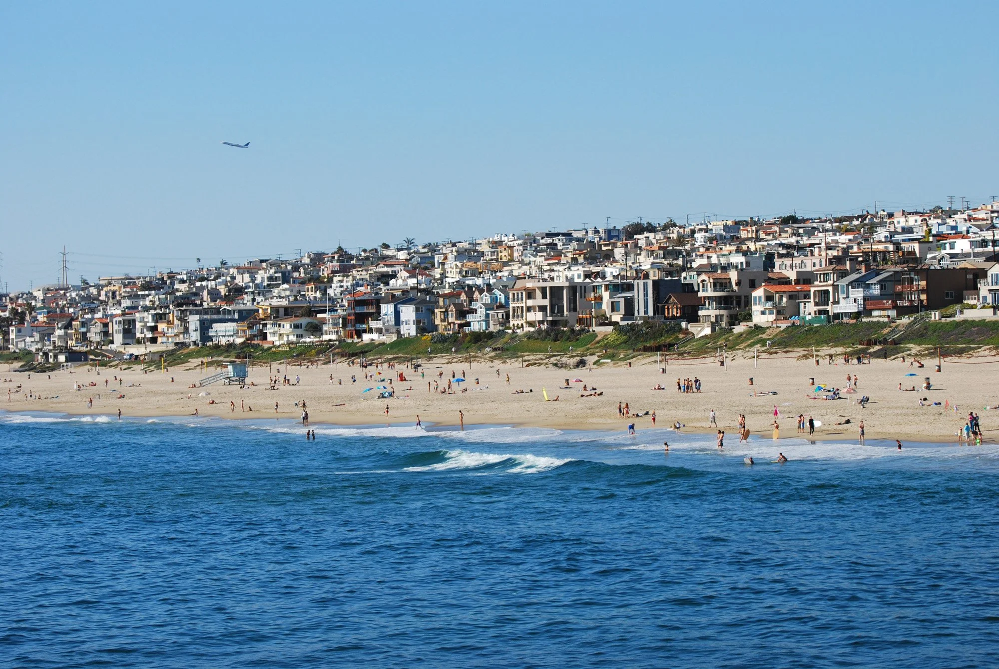 Beach scene with people enjoying the sand and waves, houses on a hill in the background, and an airplane flying in the clear blue sky.