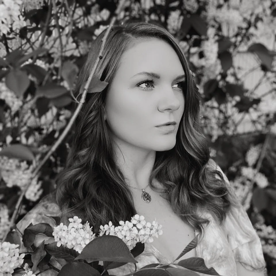 A woman with long, wavy hair and a necklace, looking to the right, surrounded by leaves and clustered flowers.