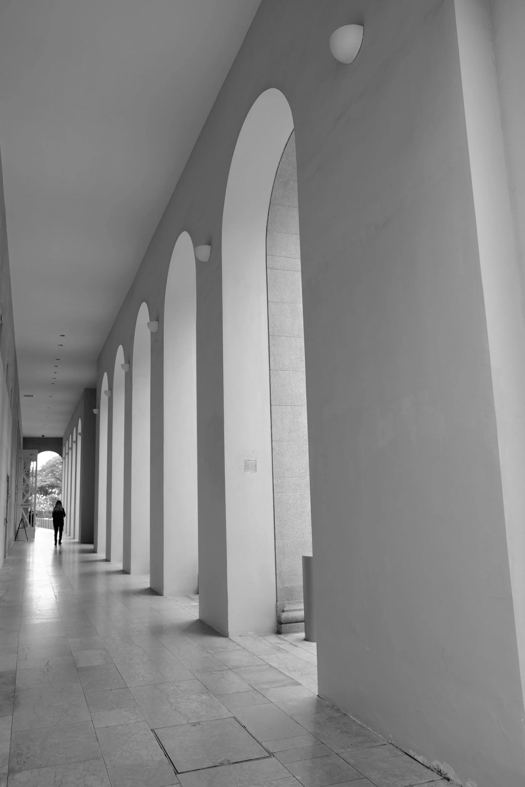 Black and white photo of a corridor with tall, arched windows and a person walking in the distance.