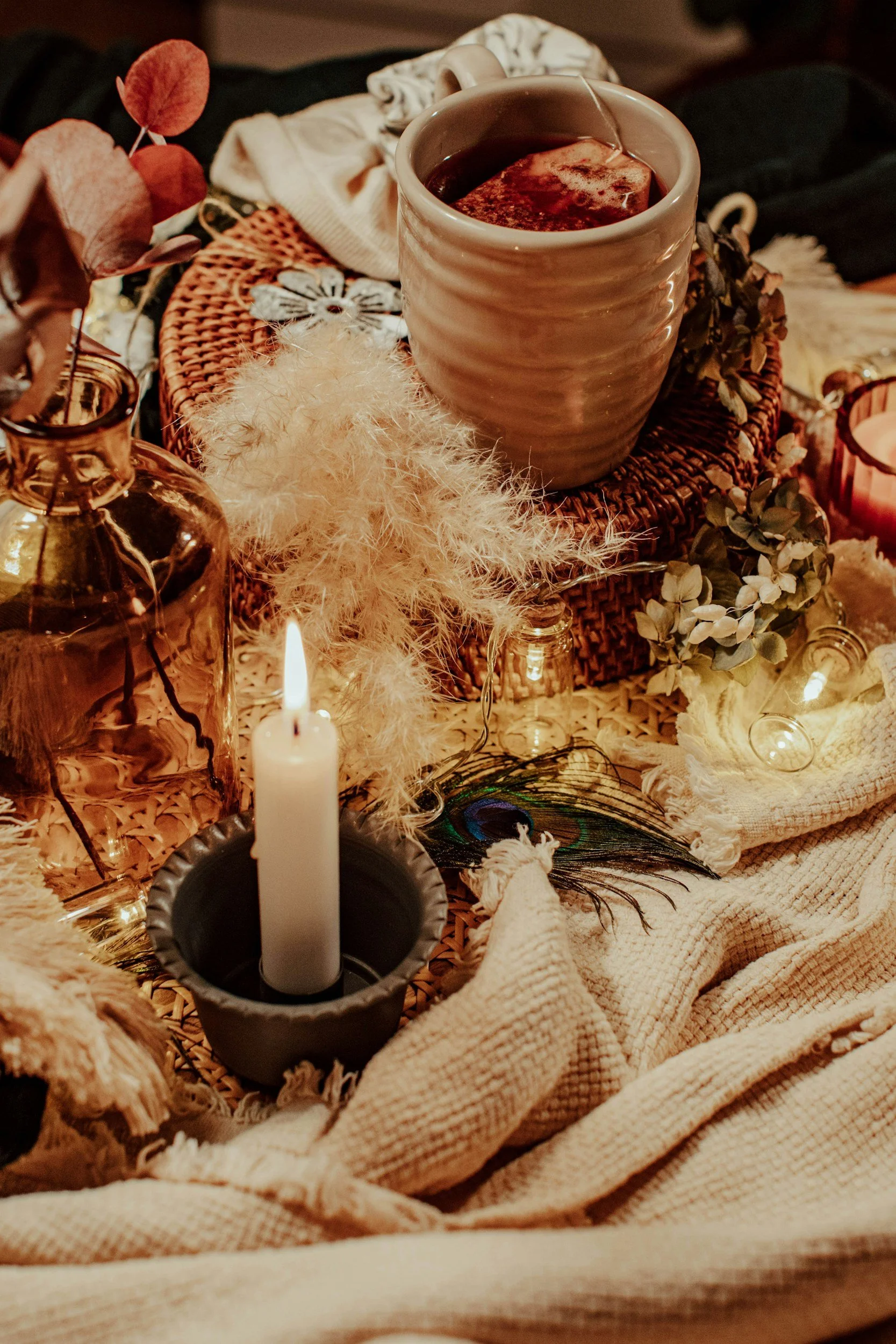 A cozy scene with a cup of hot tea or coffee, a lit white candle in a black holder, dried flowers, a peacock feather, candles, and textile layers on a woven surface.