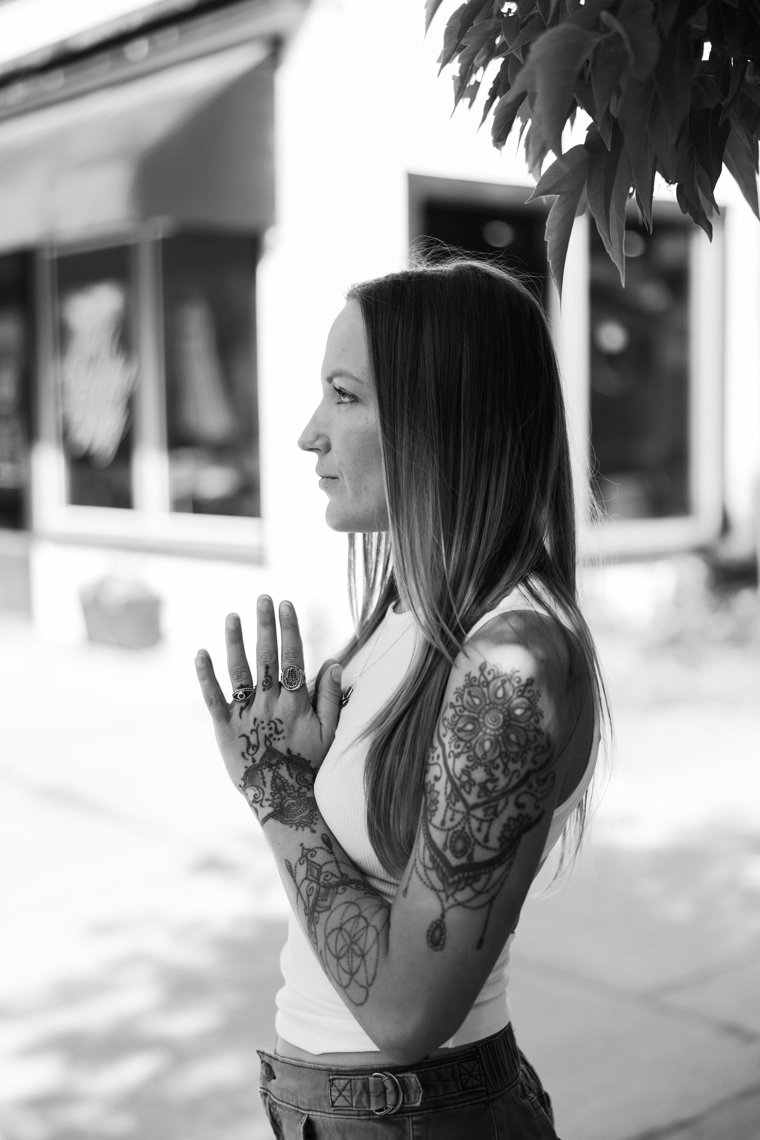 A woman with long hair and tattoos on her arms participating in a yoga or meditation practice outdoors. She has her eyes closed, hands in a prayer position, and is wearing a white tank top and dark pants. The background of the image is blurred with trees and buildings.