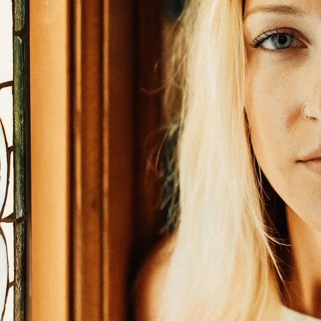 Close-up of a woman's face showing her right eye, partial nose, and blonde hair, with a wooden background and patterned window frame to the left.
