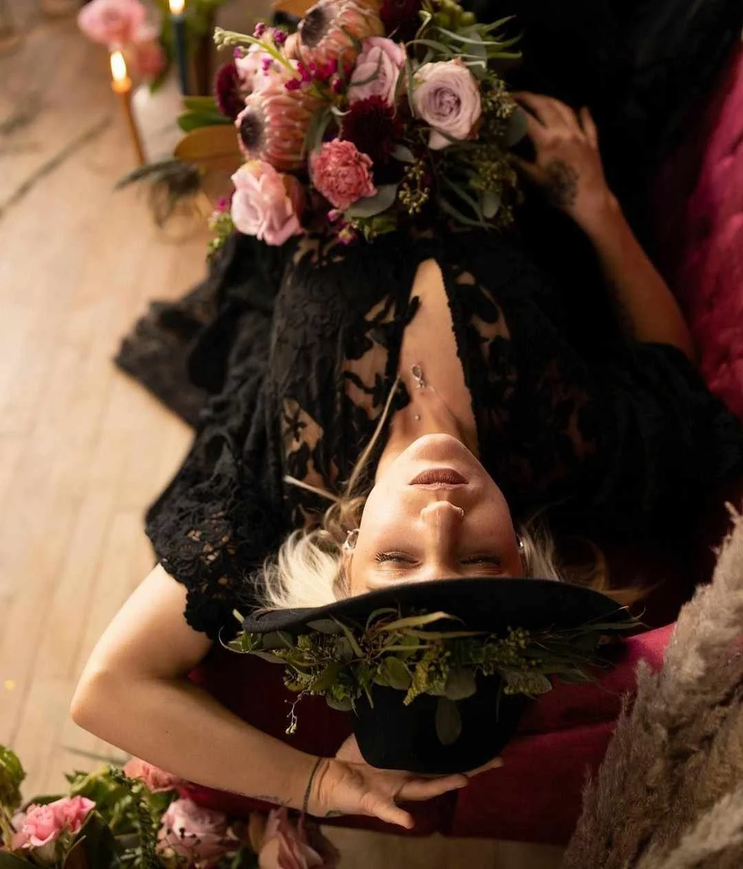 A woman with platinum blonde hair lying on her back, eyes closed, wearing a black lace dress and a black hat with a green foliage decoration. She holds a bouquet of pink and purple flowers on her chest. The background includes more flowers and warm lighting.