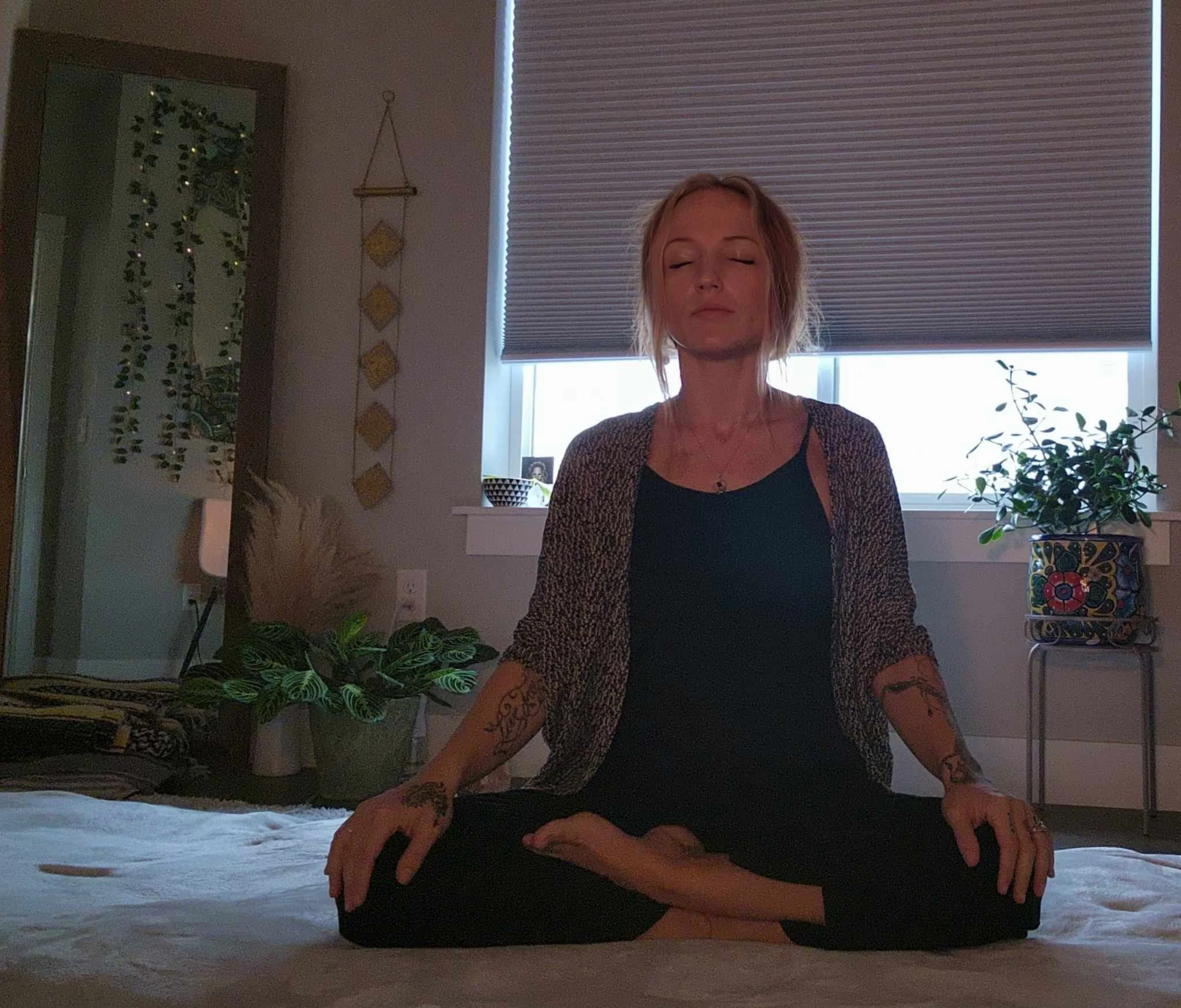 A woman practicing yoga on a bed in a bedroom, with her eyes closed and a peaceful expression, surrounded by plants and home decor.