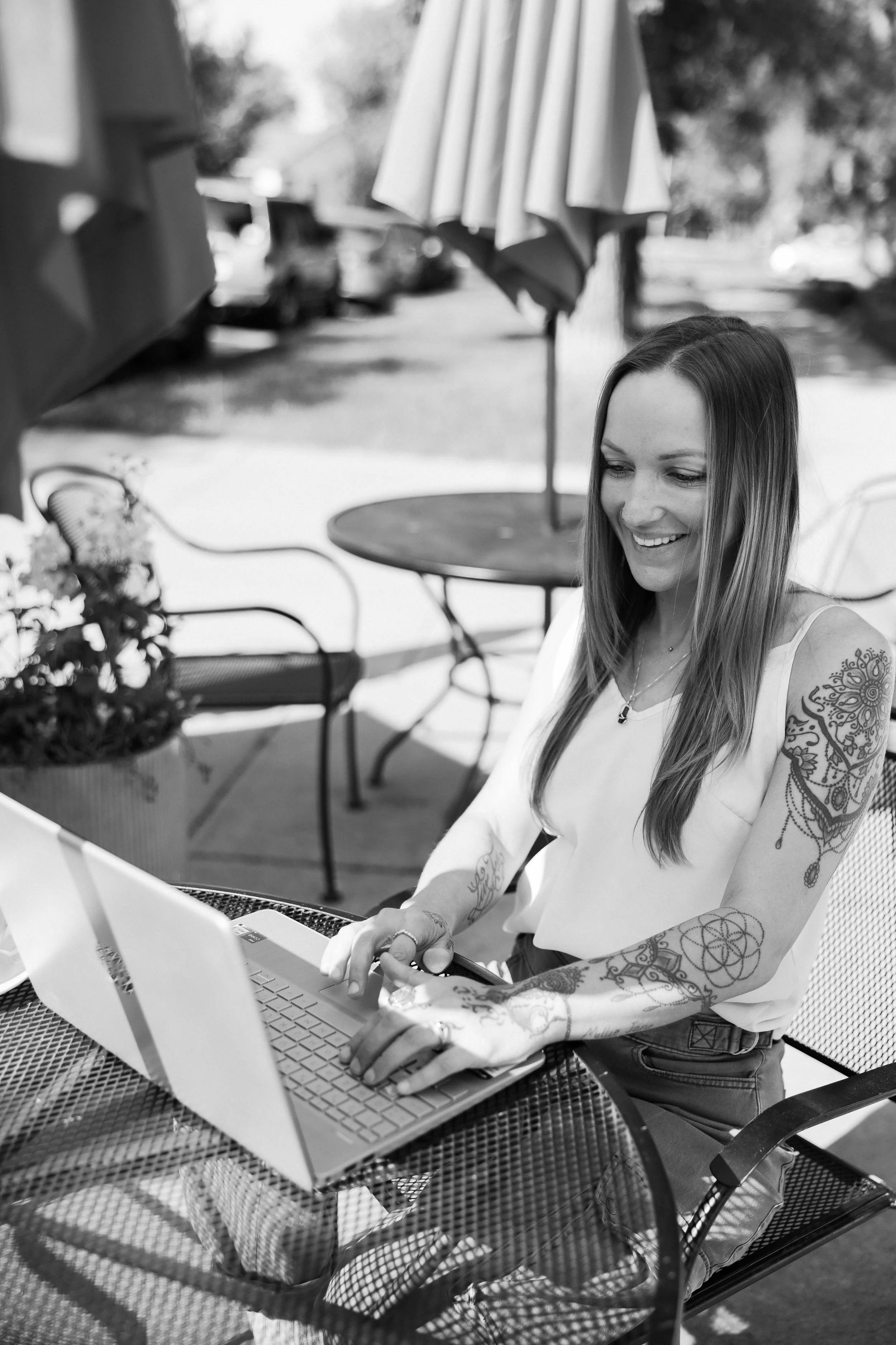 A woman with long hair and tattoos on her arms is sitting at an outdoor table, smiling while working on a laptop.