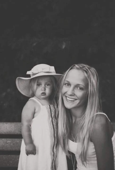 A woman and a young girl are sitting on a park bench, both smiling at the camera. The girl is wearing a large sunhat and a light-colored dress, while the woman is wearing a white top and has long blonde hair.
