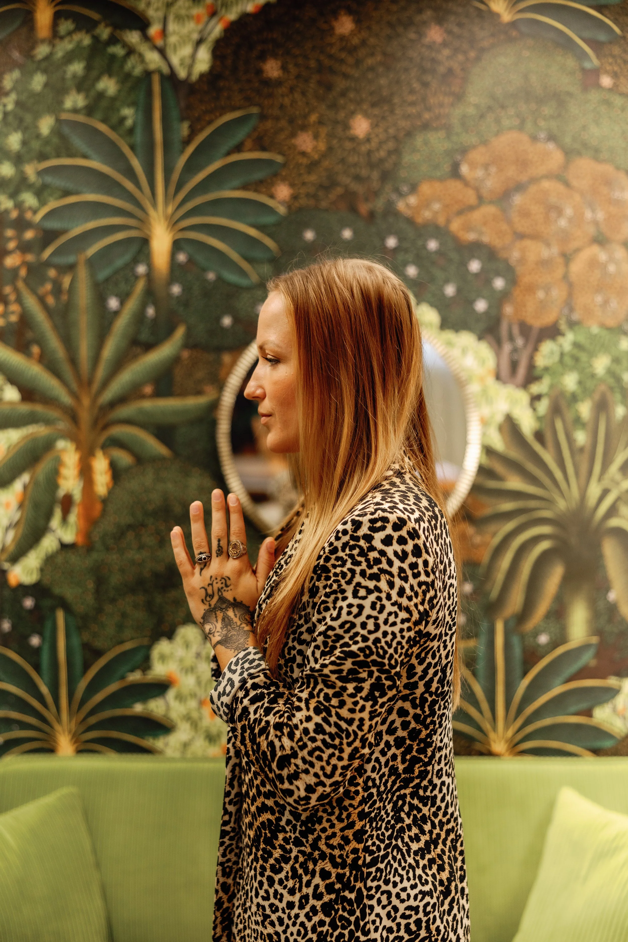 A woman with long red hair, wearing a leopard print jacket, stands with her eyes closed and hands pressed together in a prayer position against a decorative backdrop of large, green, tropical leaves and stylized orange and green trees. She has multiple rings and a tattoo on her left hand.