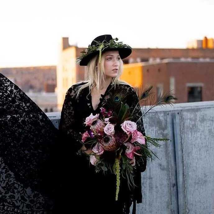 A young woman dressed in black with a wide-brimmed black hat decorated with greenery, holding a bouquet of pink roses, peacock feathers, and greenery, standing outdoors on a rooftop with city buildings in the background.