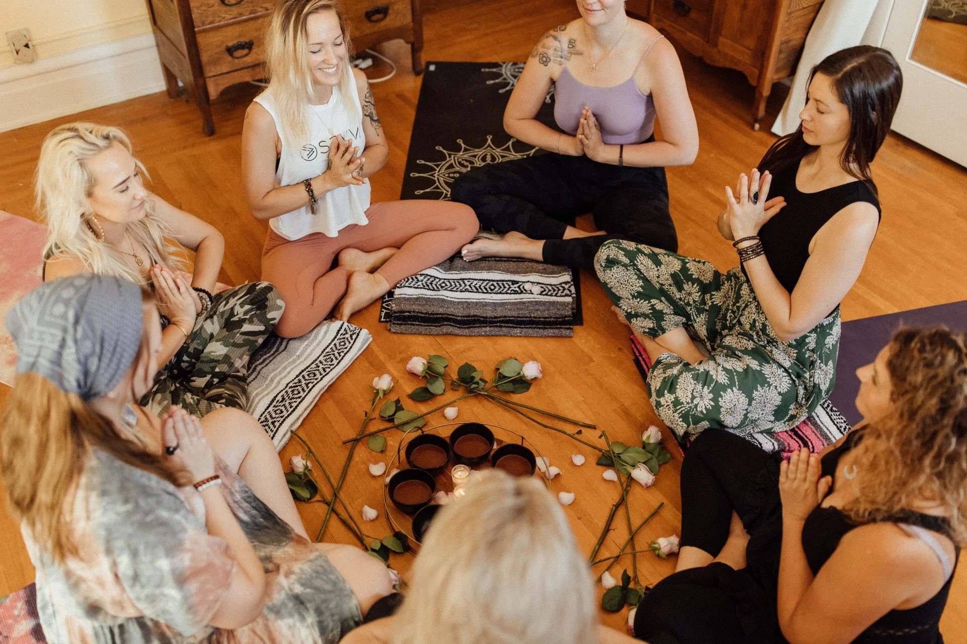A group of women sitting in a circle on the floor with roses, candles, and candles, engaging in a meditation or prayer circle in a cozy room with wooden floors and furniture.
