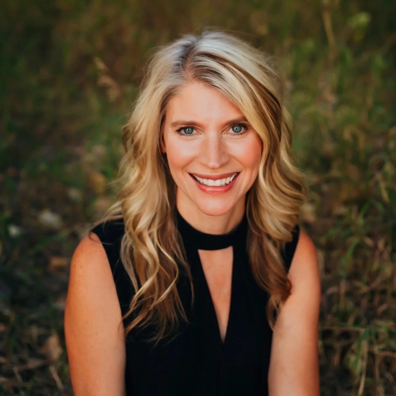 A woman with blonde hair and blue eyes smiling outdoors, wearing a black sleeveless top.