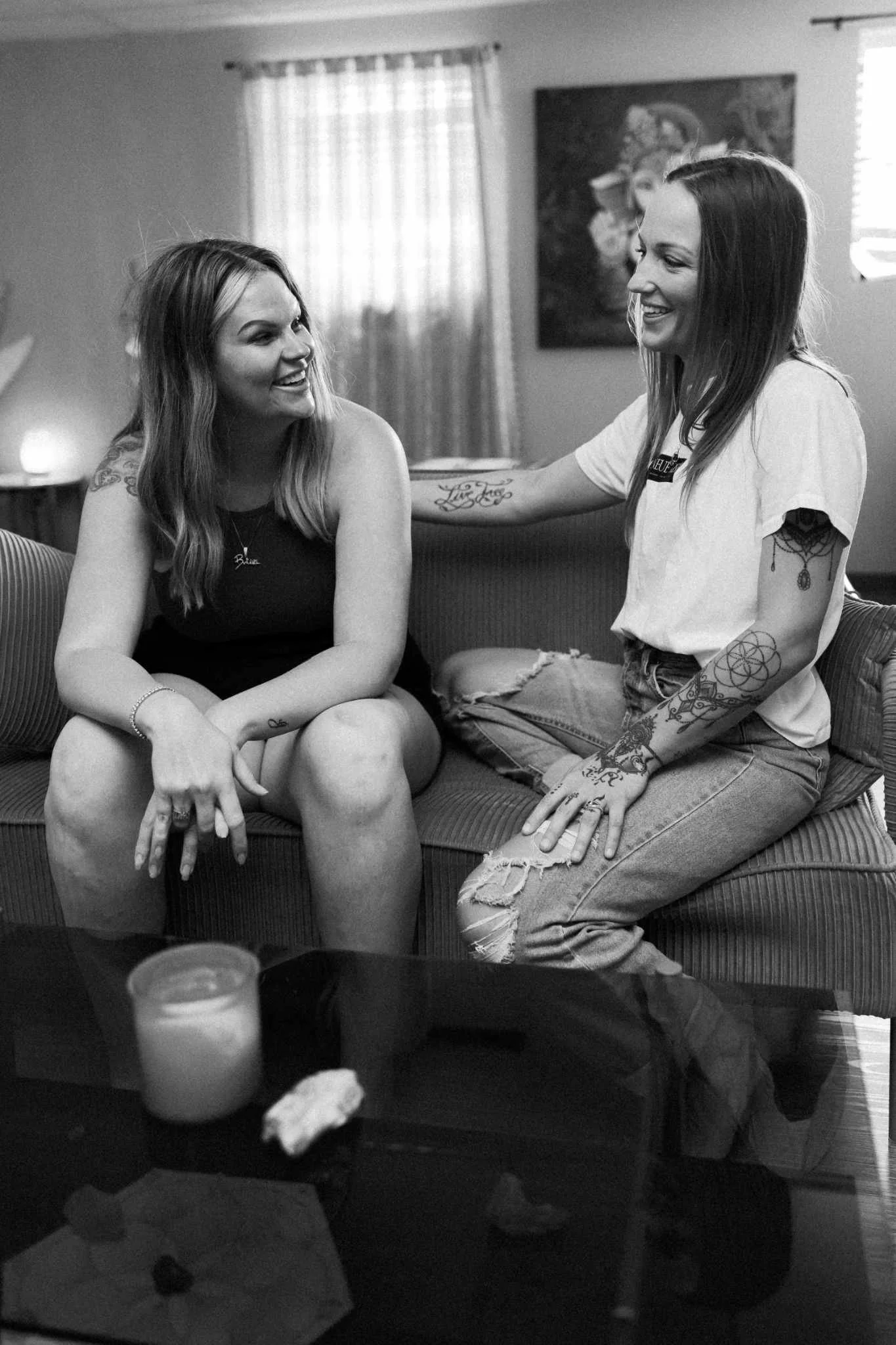 Two women smiling and talking to each other in a living room, sitting on a couch. One woman has long hair, wearing a tank top, and the other has shoulder-length hair, wearing a t-shirt and ripped jeans. There are candles, a picture on the wall, and a window with curtains in the background.