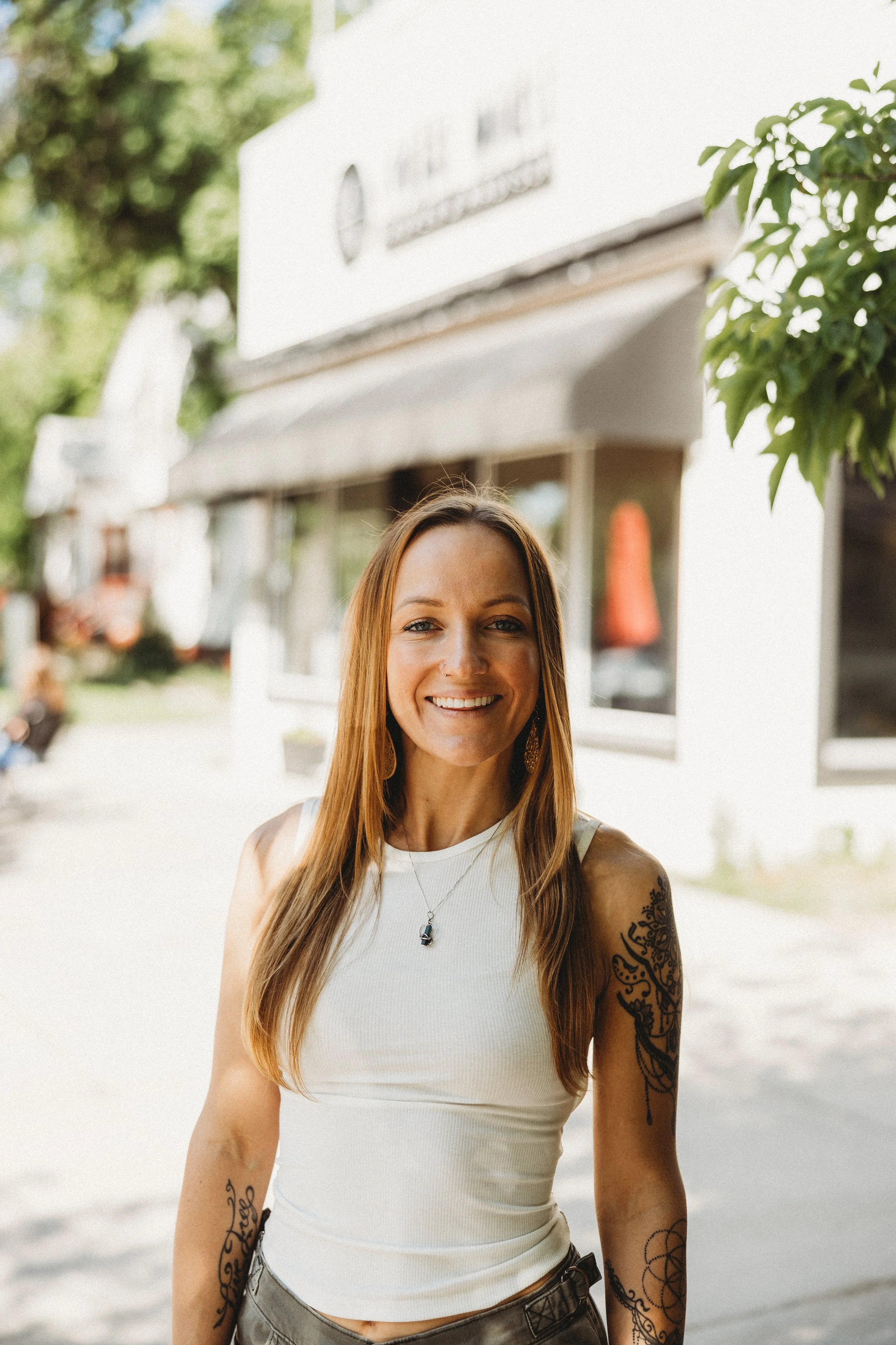 A smiling woman with long hair, tattoos on her arms, wearing a white sleeveless top and a necklace, standing outside on a sunny day with a blurred background of trees and buildings.