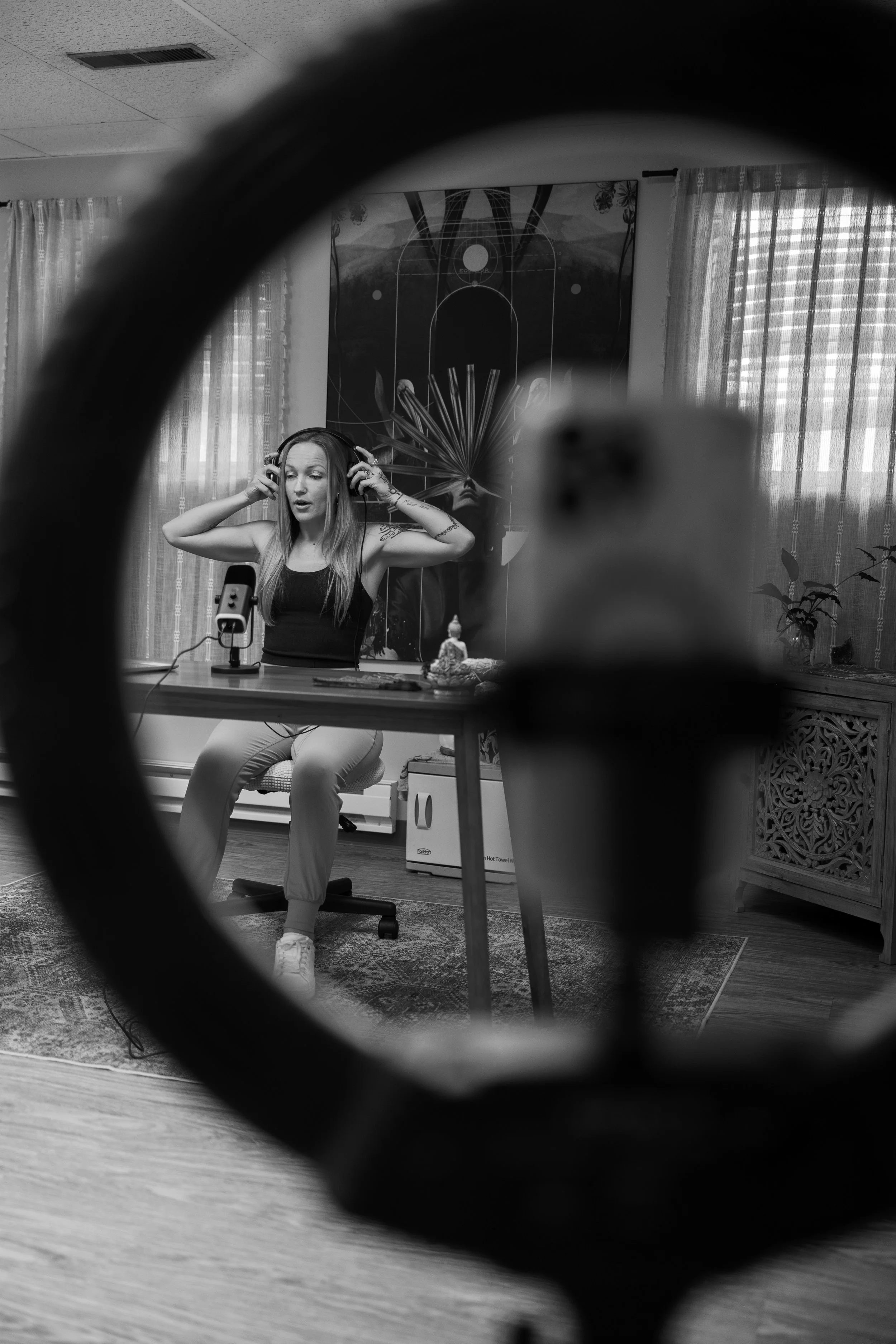 Black and white photo of a woman sitting in a room during a podcast recording session, seen through a circular frame. She is wearing headphones, sitting at a table with a microphone, and appears to be speaking or singing. The room has curtains and decorative artwork on the wall.