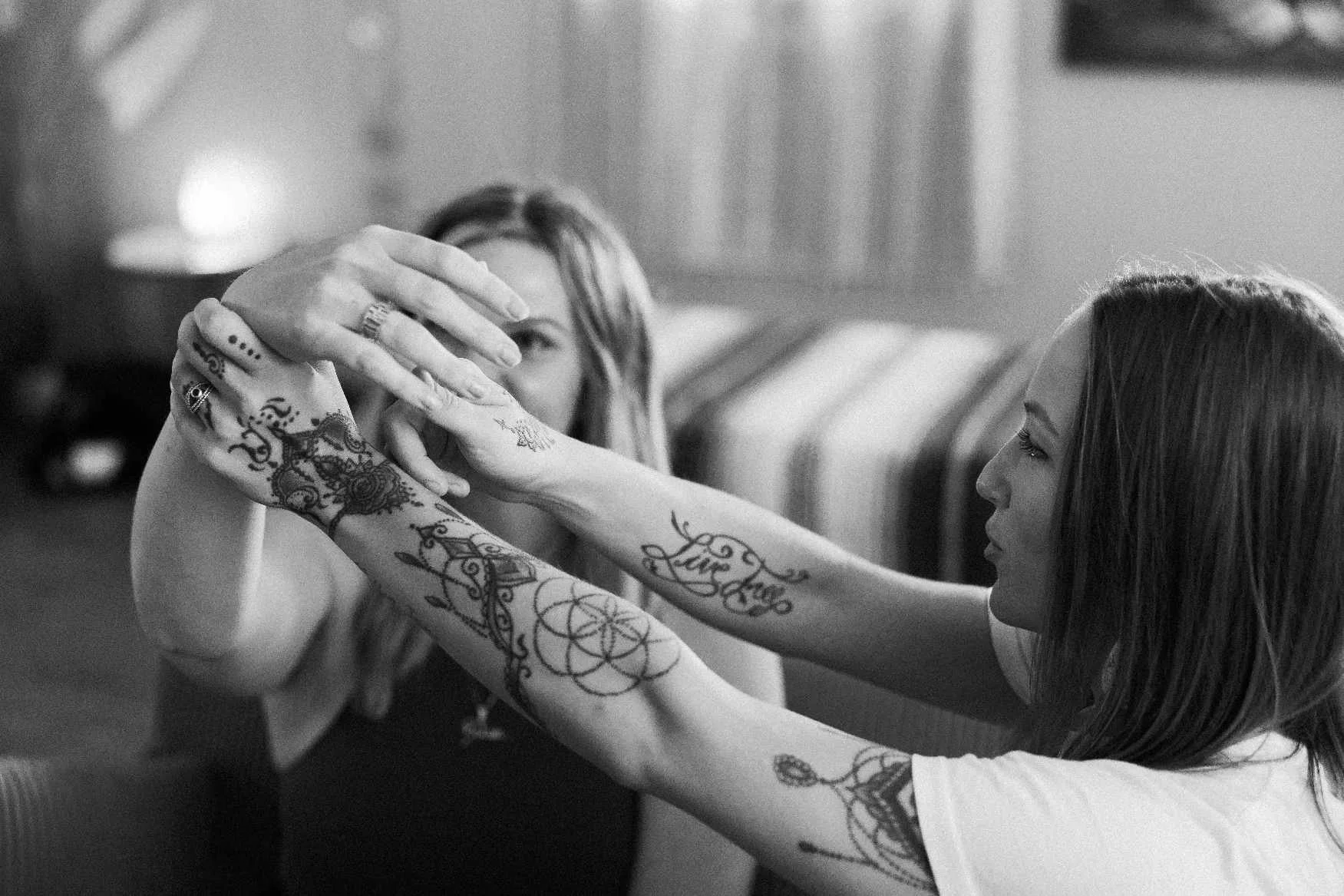 Two women with tattoos on their arms are holding hands, their faces close together, in a black-and-white photo.