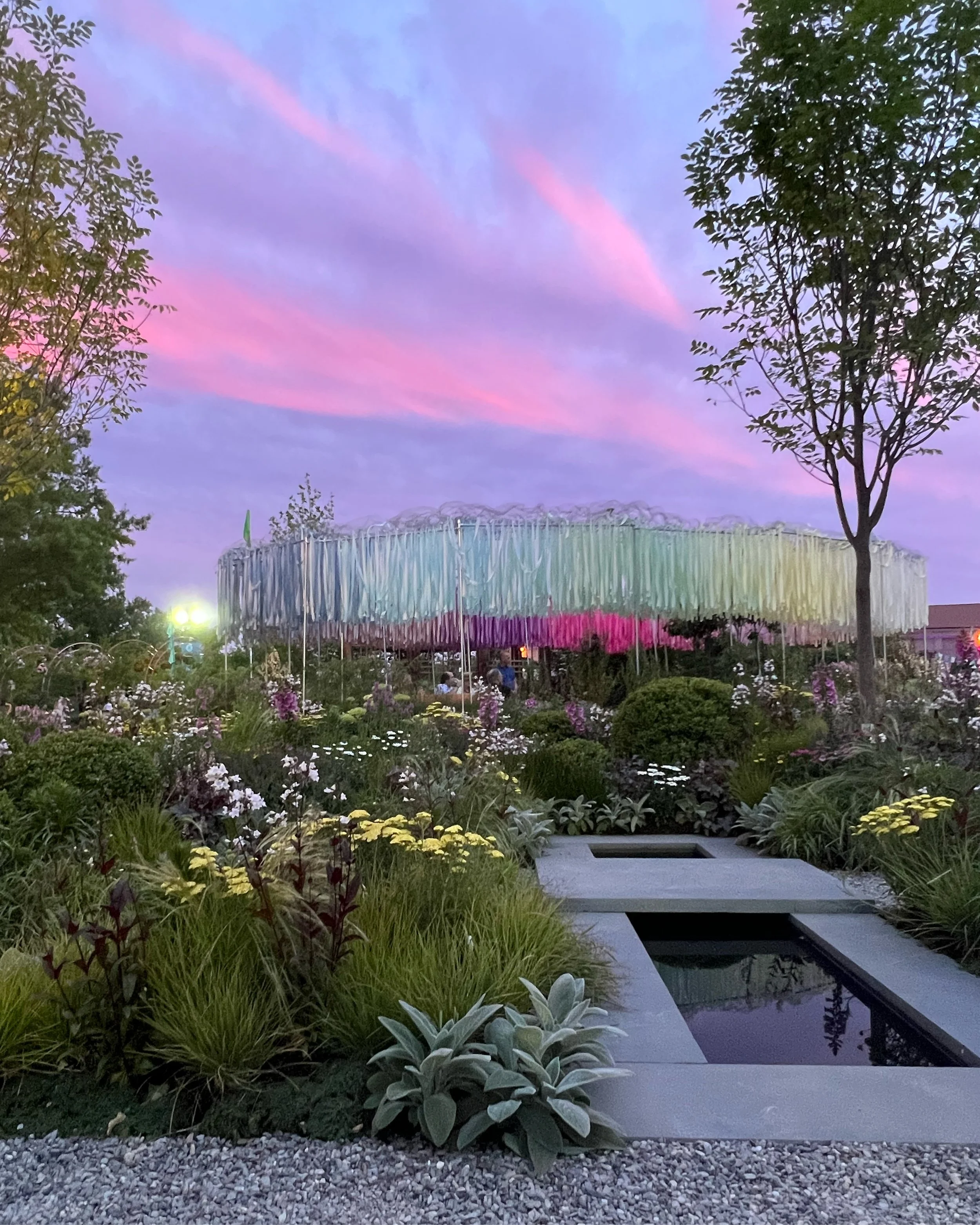 A colorful outdoor art installation surrounded by a garden with various flowers and plants during twilight, with a vibrant pink and purple sky above.