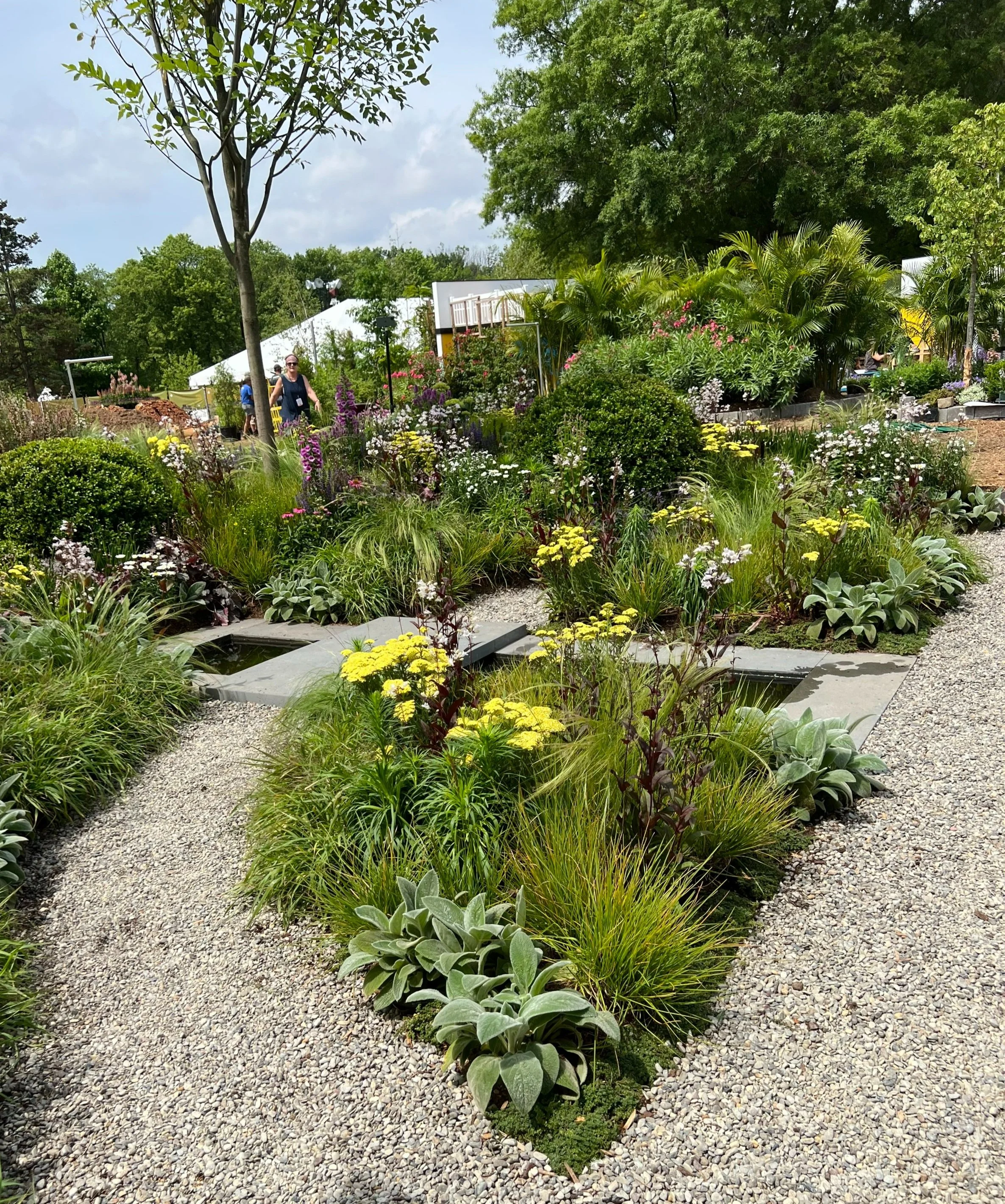 A vibrant garden with various green plants and colorful flowers, a gravel pathway, and a person walking amidst the greenery on a partly cloudy day.