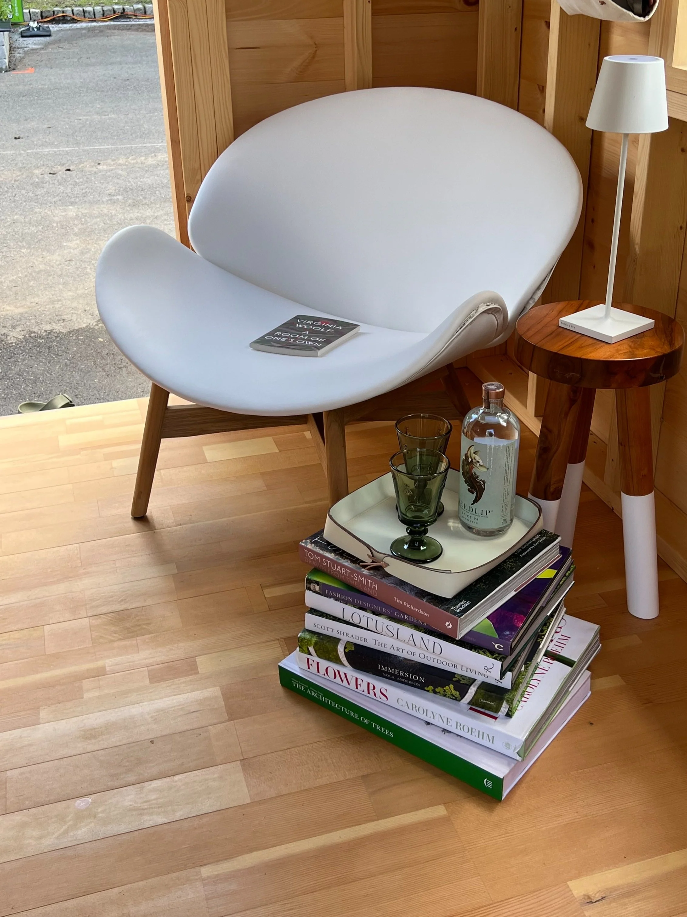 A modern white lounge chair with wooden legs, a small round wooden side table with a white lamp, and a stack of books topped with a tray holding three glasses and a bottle, set on a wooden floor near a window.