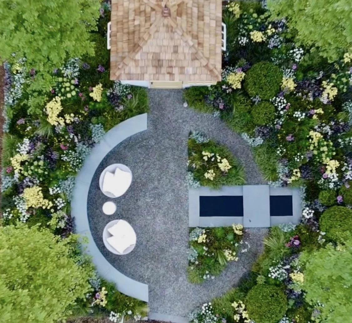 Aerial view of a garden with a gravel pathway, surrounded by colorful flowers and greenery, featuring a small wooden shed on one side and two white chairs with cushions on the other.