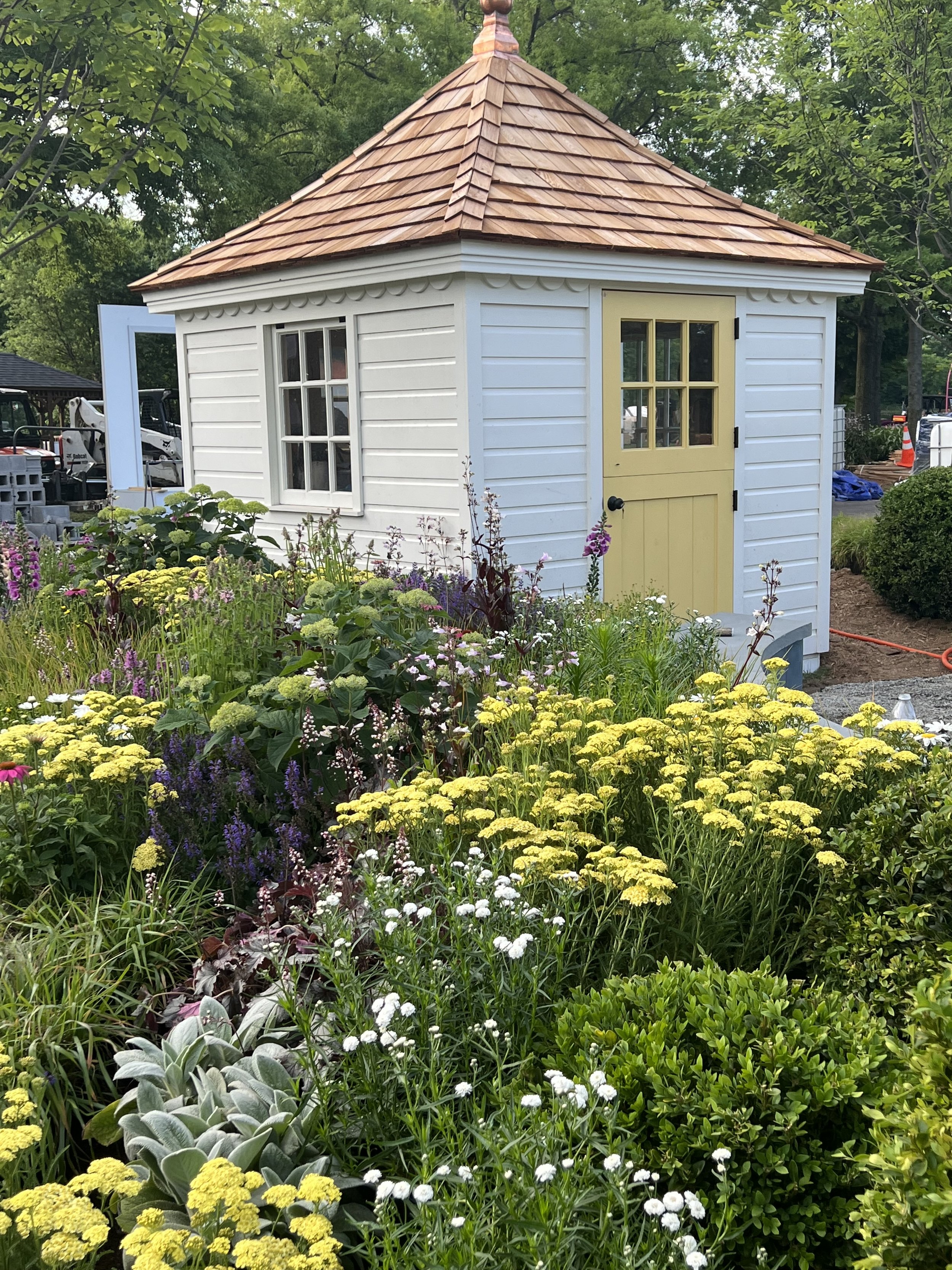 A small white garden shed with a yellow door and a shingled, pyramid-shaped roof, surrounded by colorful flowers and greenery.