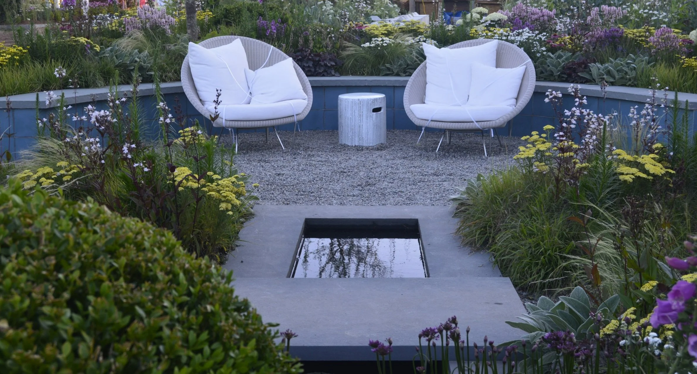 Two white modern outdoor chairs with cushions facing each other, separated by a small round white table, set in a garden with various flowering plants, and a small water feature with a reflective surface in the center.