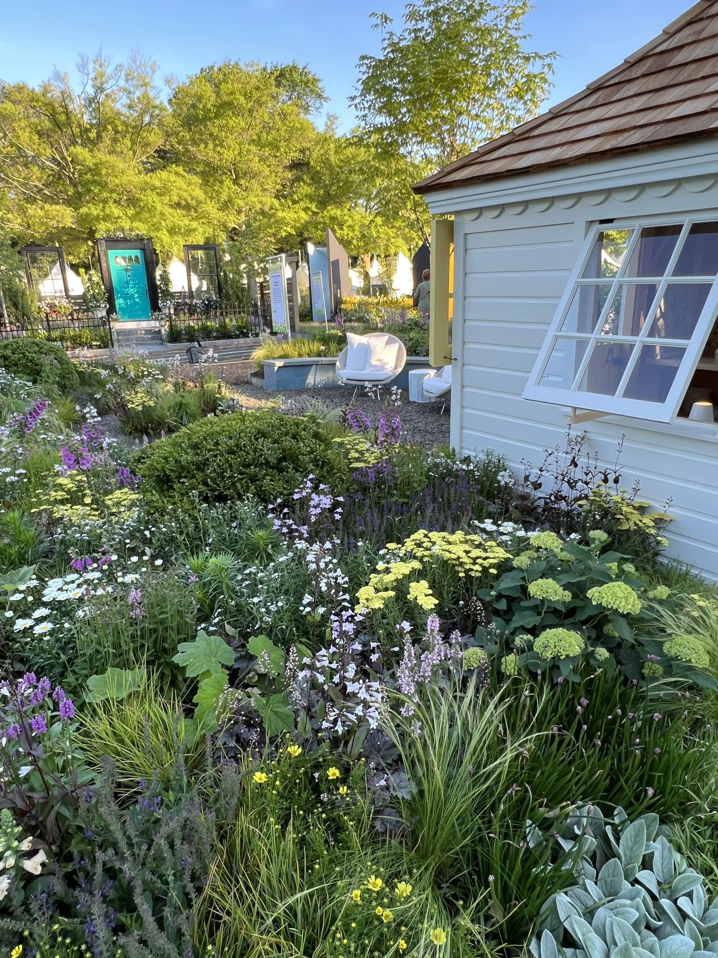 A lush garden with colorful flowers and green foliage next to a white building with a slightly open window, in a setting with decorative objects, outdoor seating, and informational signs.