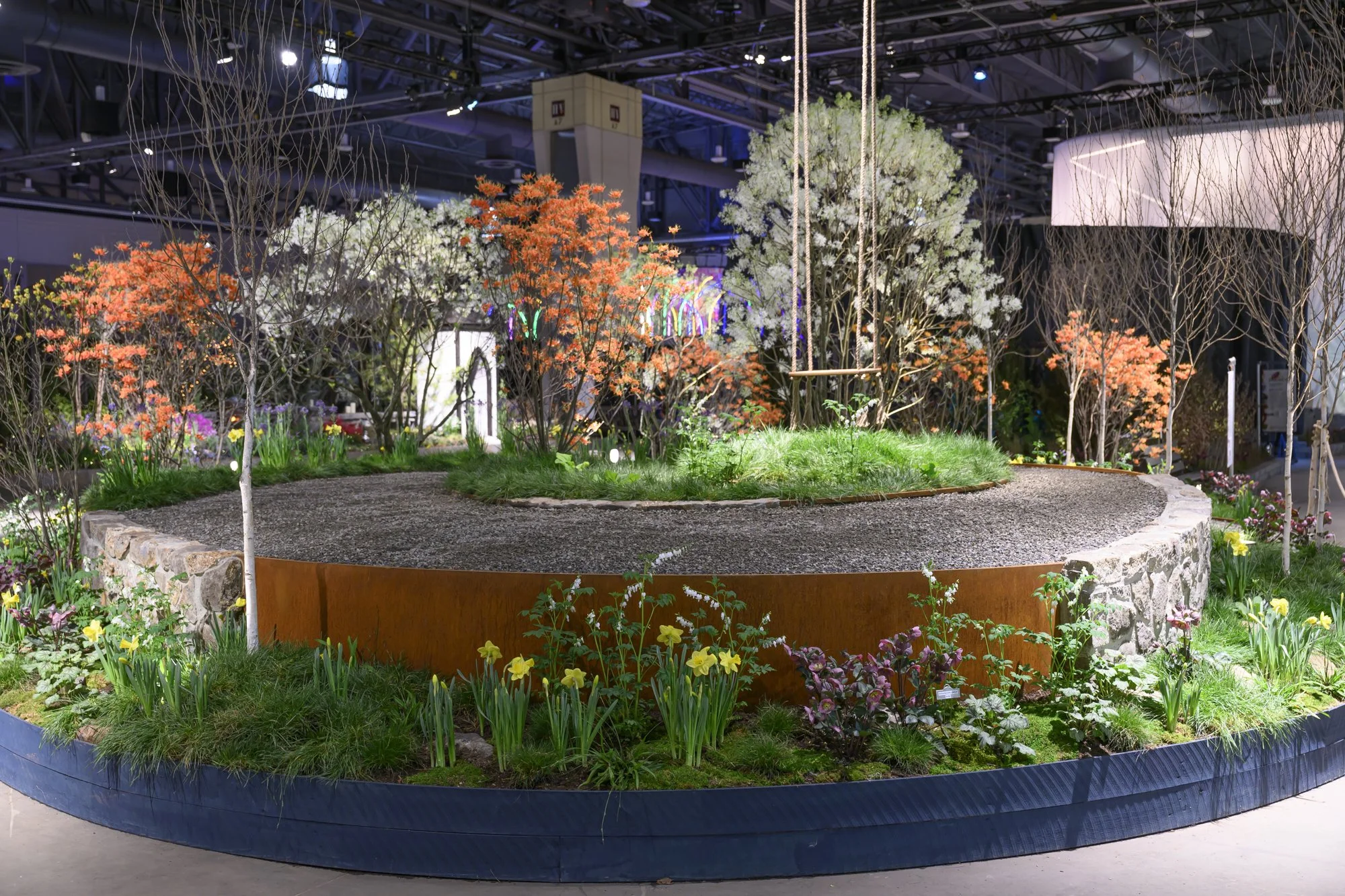 Indoor garden exhibit featuring a circular raised garden bed with various flowering trees, shrubs, and plants, illuminated with spotlights in a modern setting.