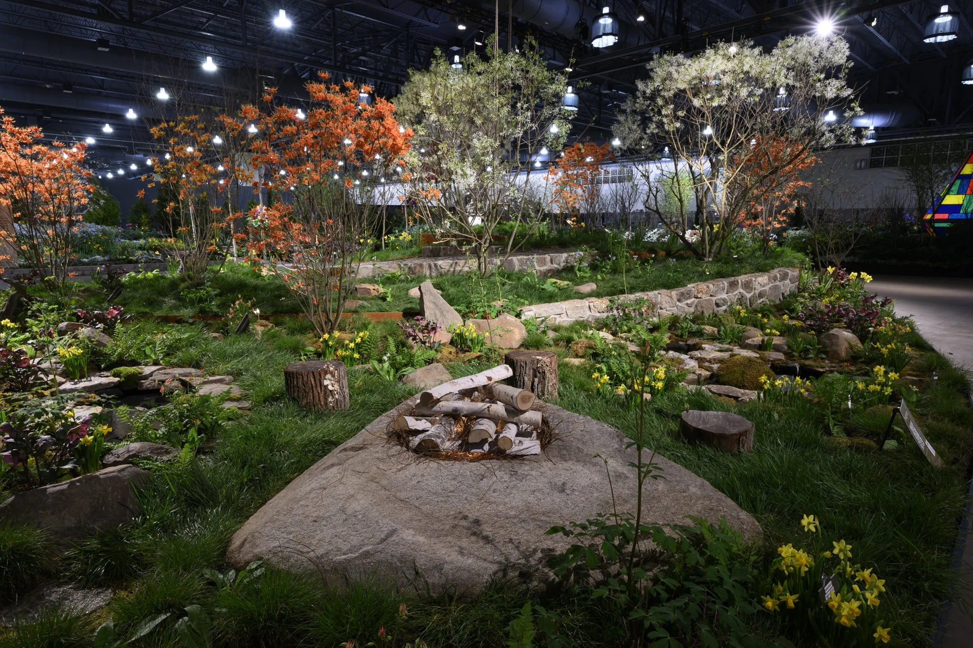 Indoor garden with various plants, trees, and flowers illuminated by bright ceiling lights. Rocks and logs are used as decoration, and a stone path winds through the garden.