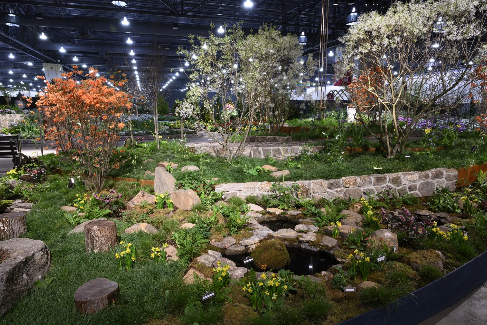 Indoor garden exhibit with trees, shrubs, flowers, rocks, and small pond, under bright artificial lighting.