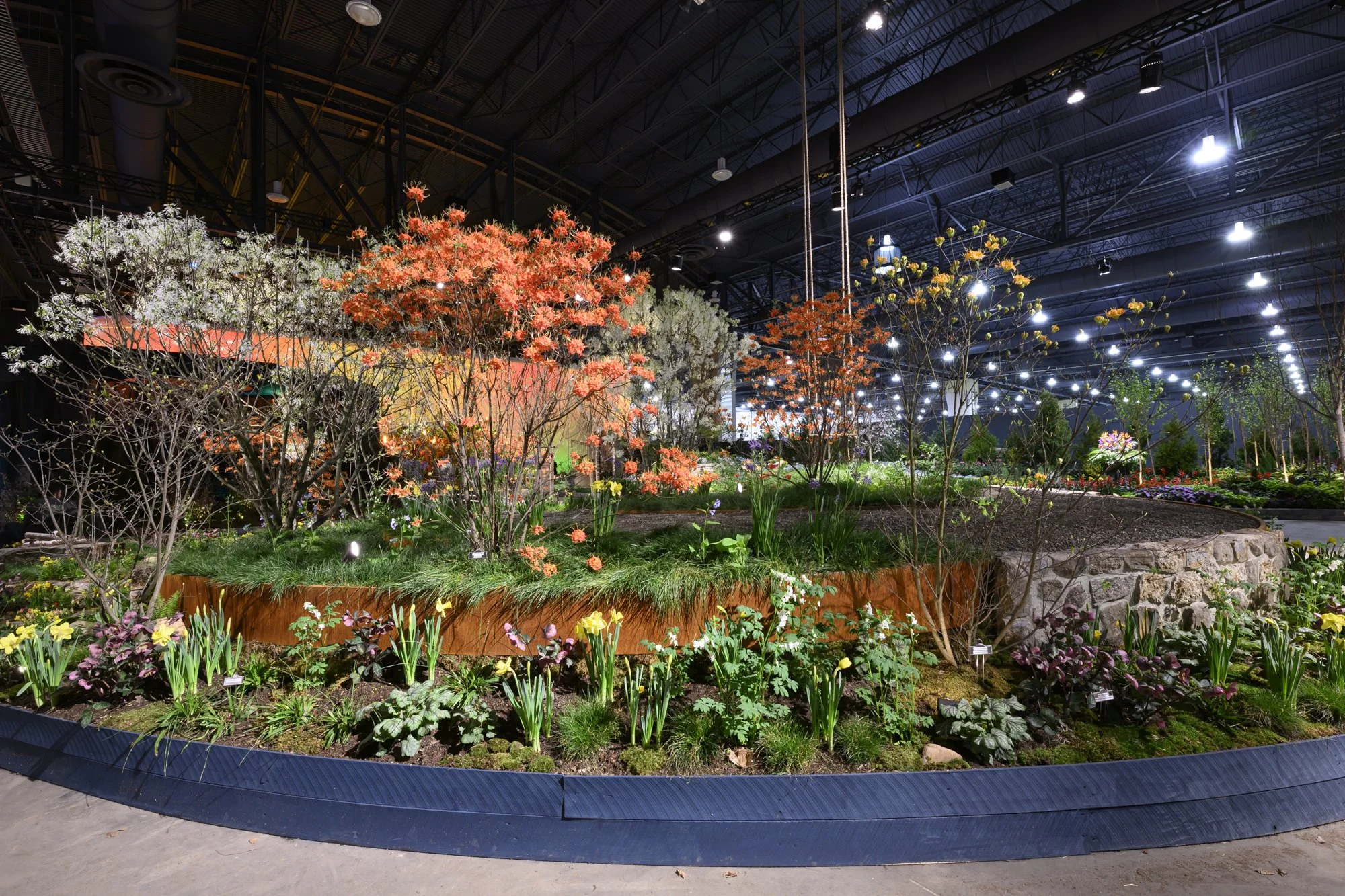 Indoor display of colorful flowering trees and plants in a botanical garden exhibit with overhead lights and dark industrial ceiling.