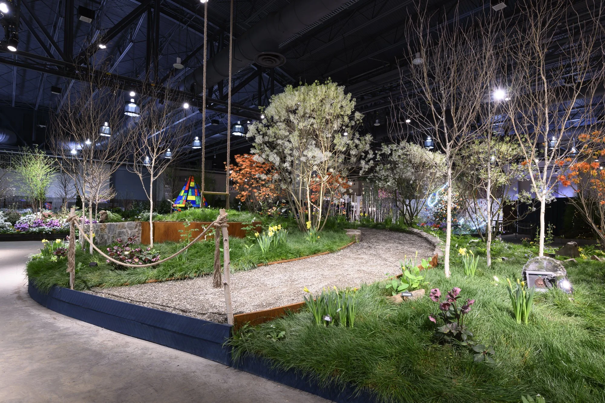 Indoor garden display with various trees, flowering plants, and a pathway, illuminated by overhead lighting.