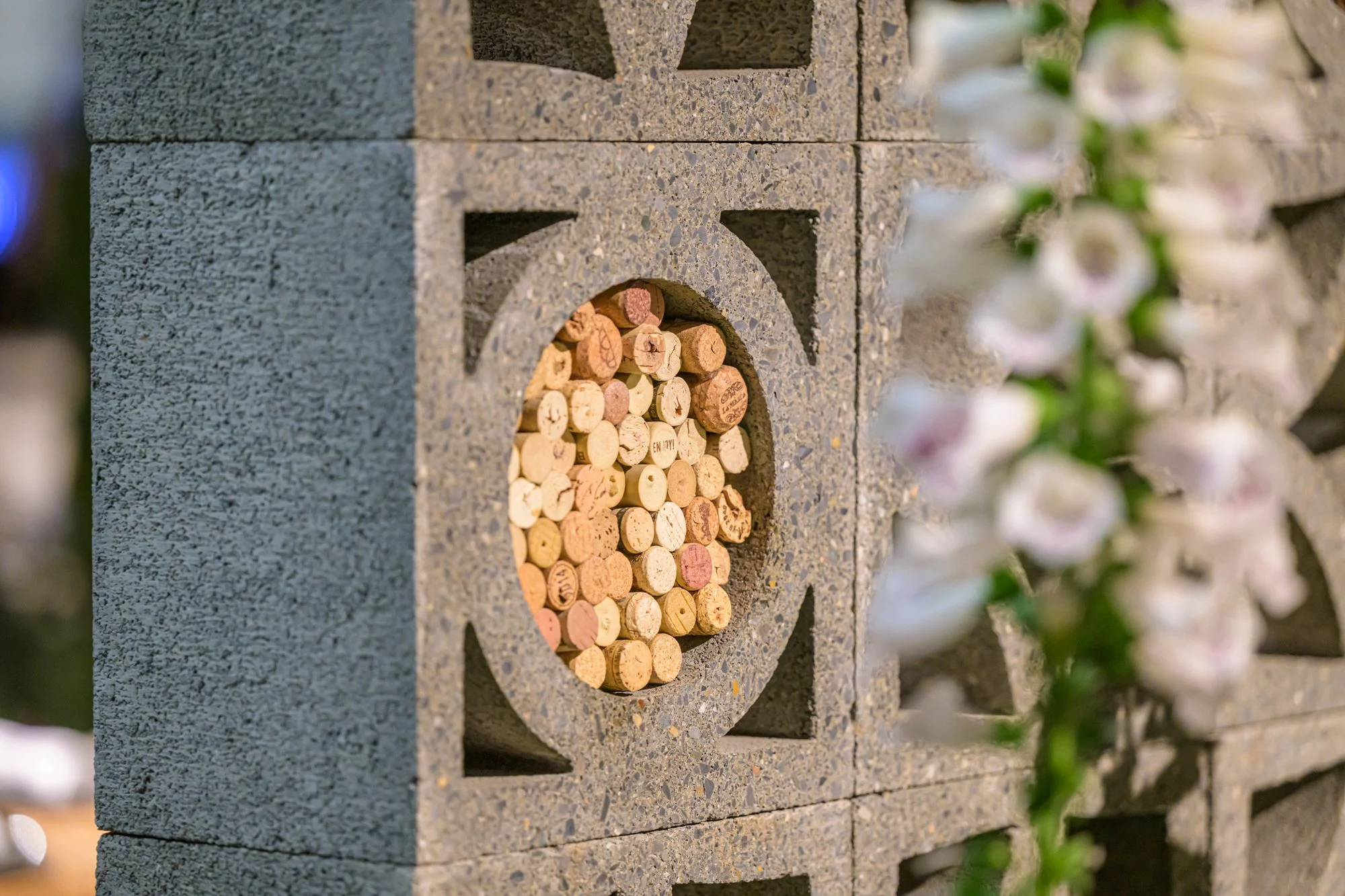 A decorative structure made of gray concrete bricks with a circular opening filled with wine corks, surrounded by blurred pink and white flowers on the right side.