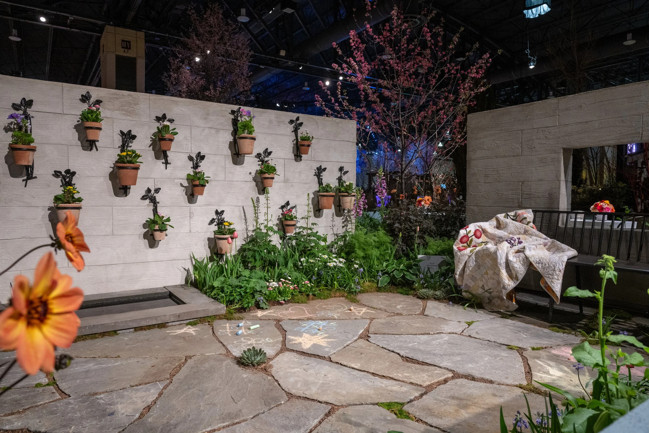 Indoor garden with potted plants on a white wall, chalk-drawn games on stone pathway, and pink flowering trees in the background.