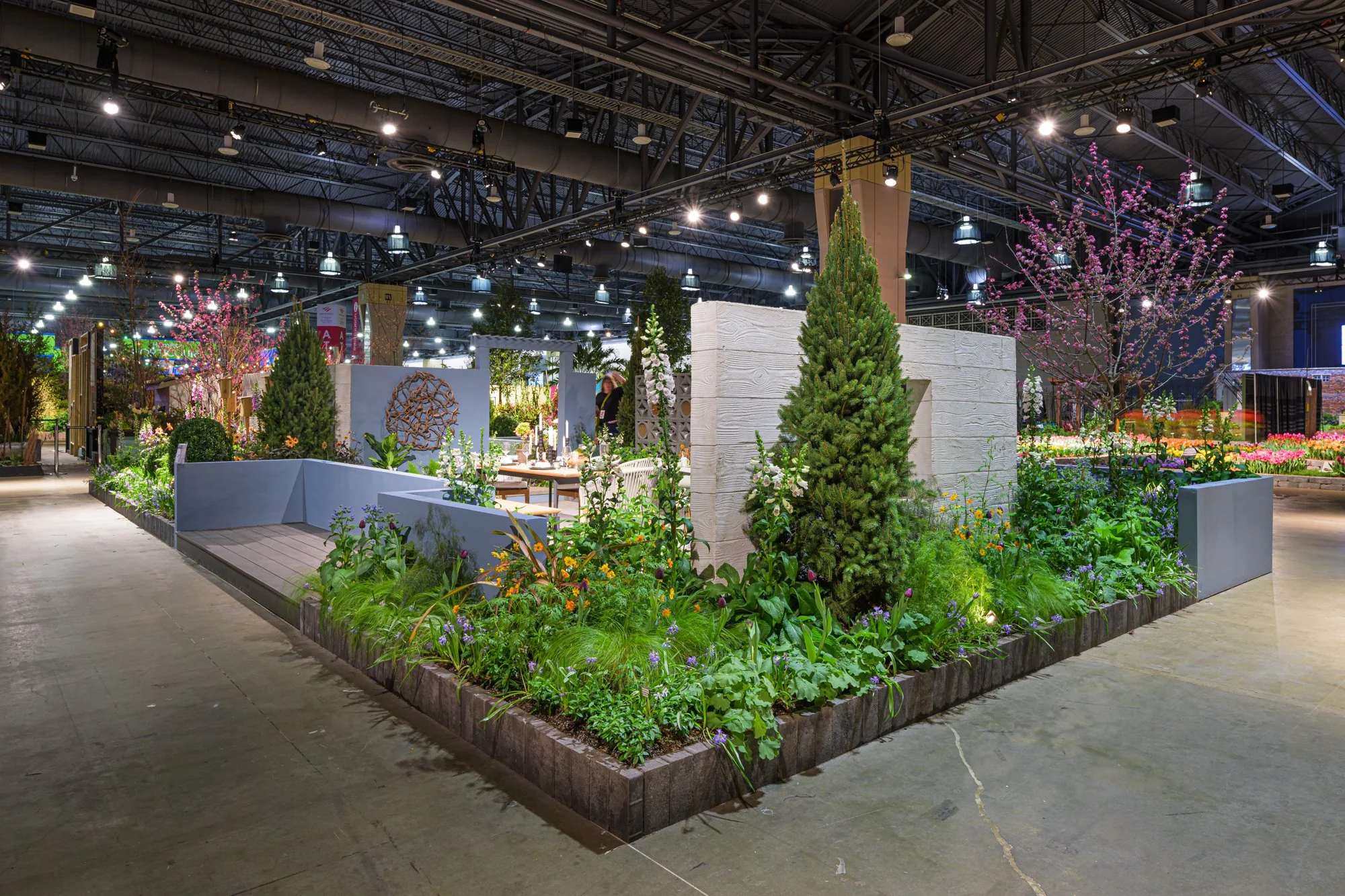 Indoor garden display with various trees, plants, and flowers, including a tall pine tree and cherry blossoms, in a spacious exhibition hall with industrial ceiling and lighting.