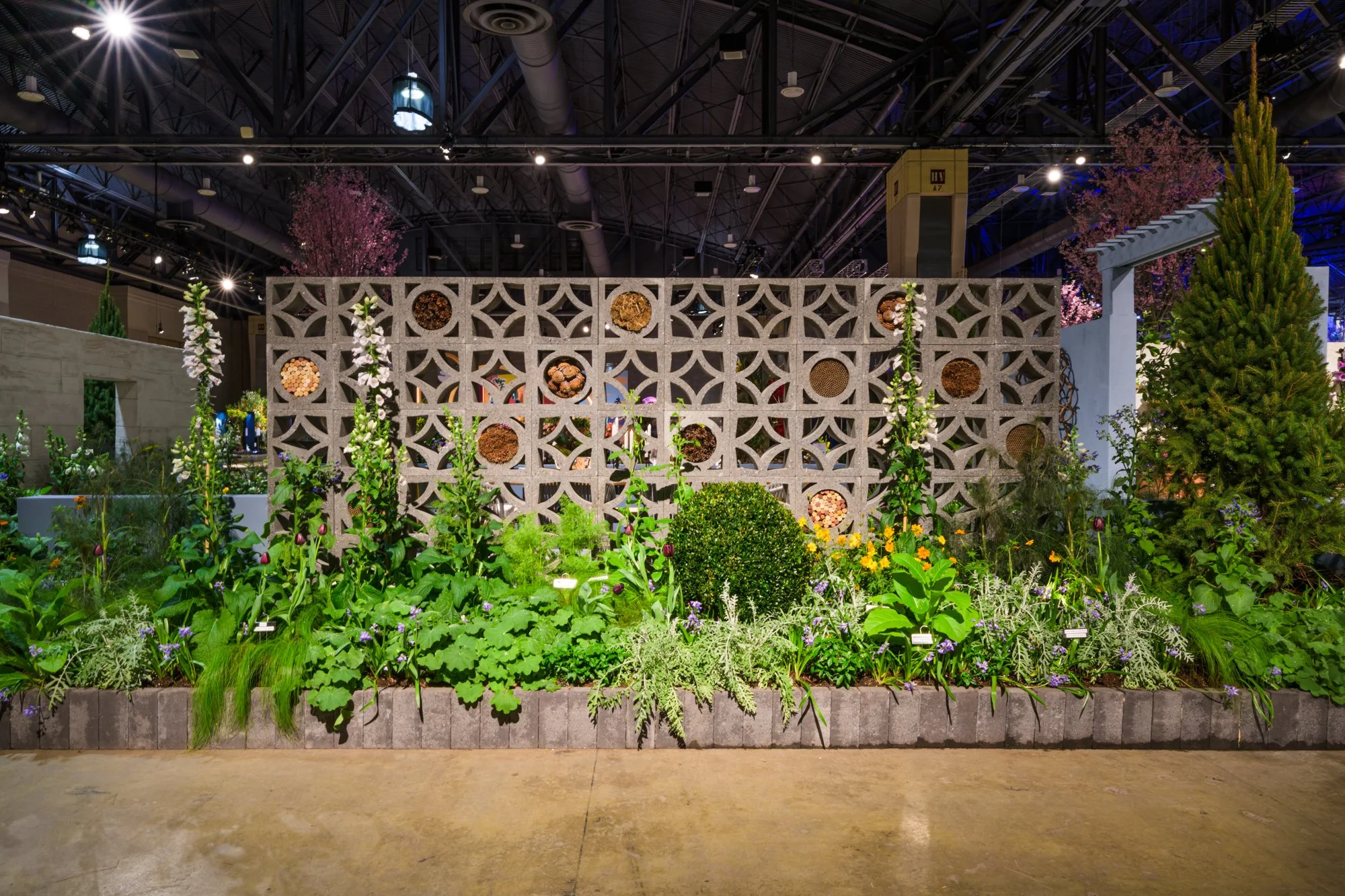 Indoor garden with a decorative concrete block wall, various green plants, and trees in an exhibition hall.