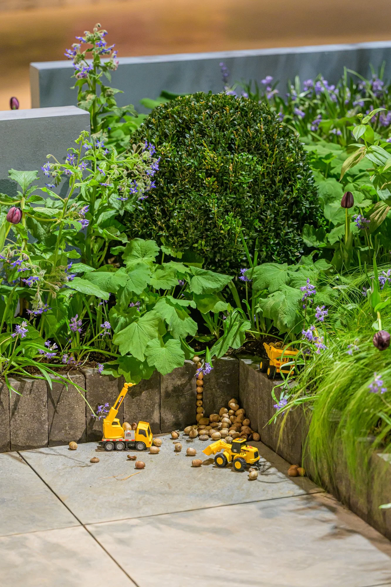Miniature construction toy trucks on a patio among green plants and purple flowers.