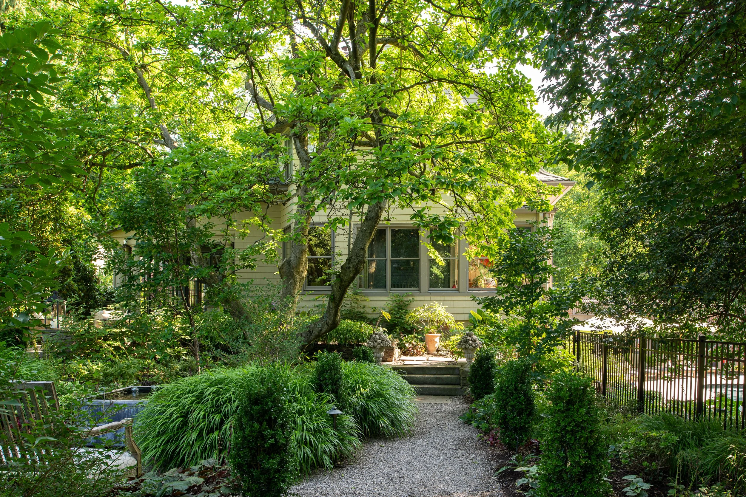 A lush green garden with a gravel path leading to a house with large windows, surrounded by trees, shrubs, and decorative plants.