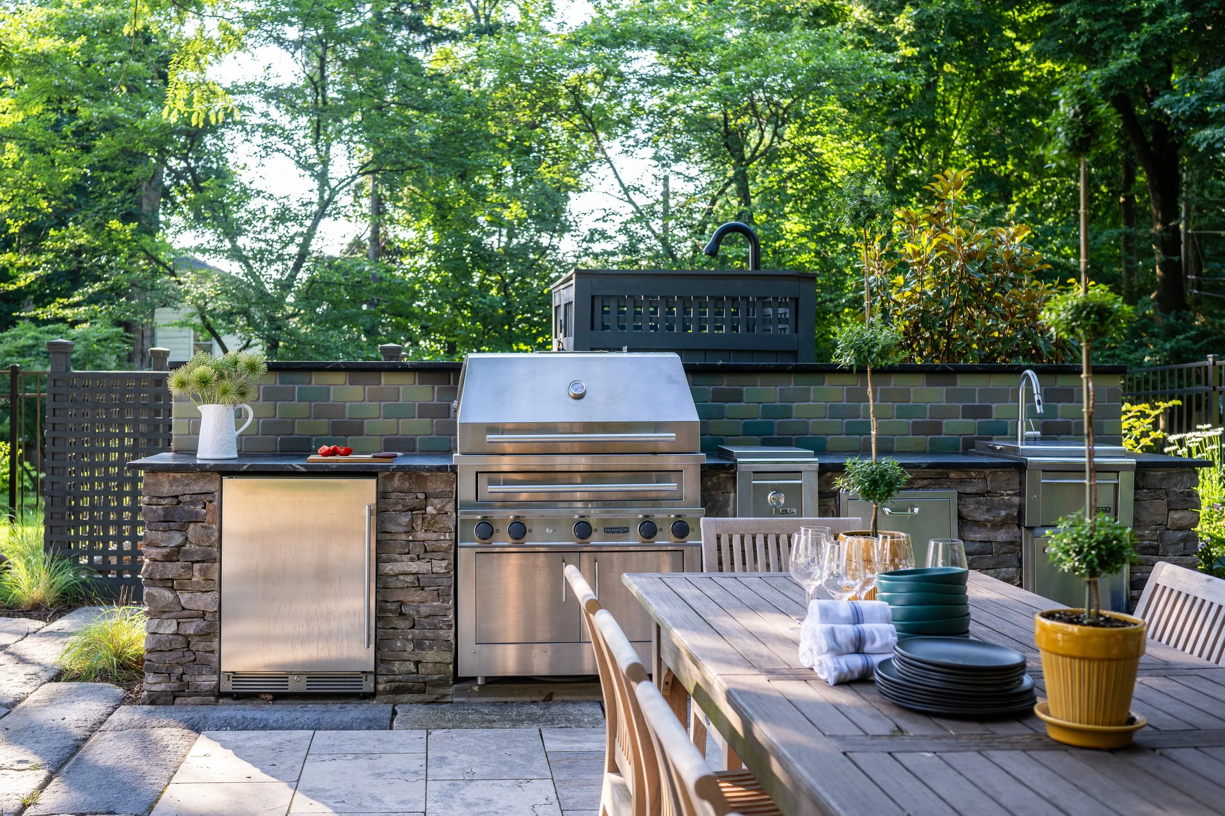 Outdoor kitchen with stainless steel grill, sink, and stone countertop, surrounded by a dining table with plates, glasses, and potted plants, set in a lush green backyard.