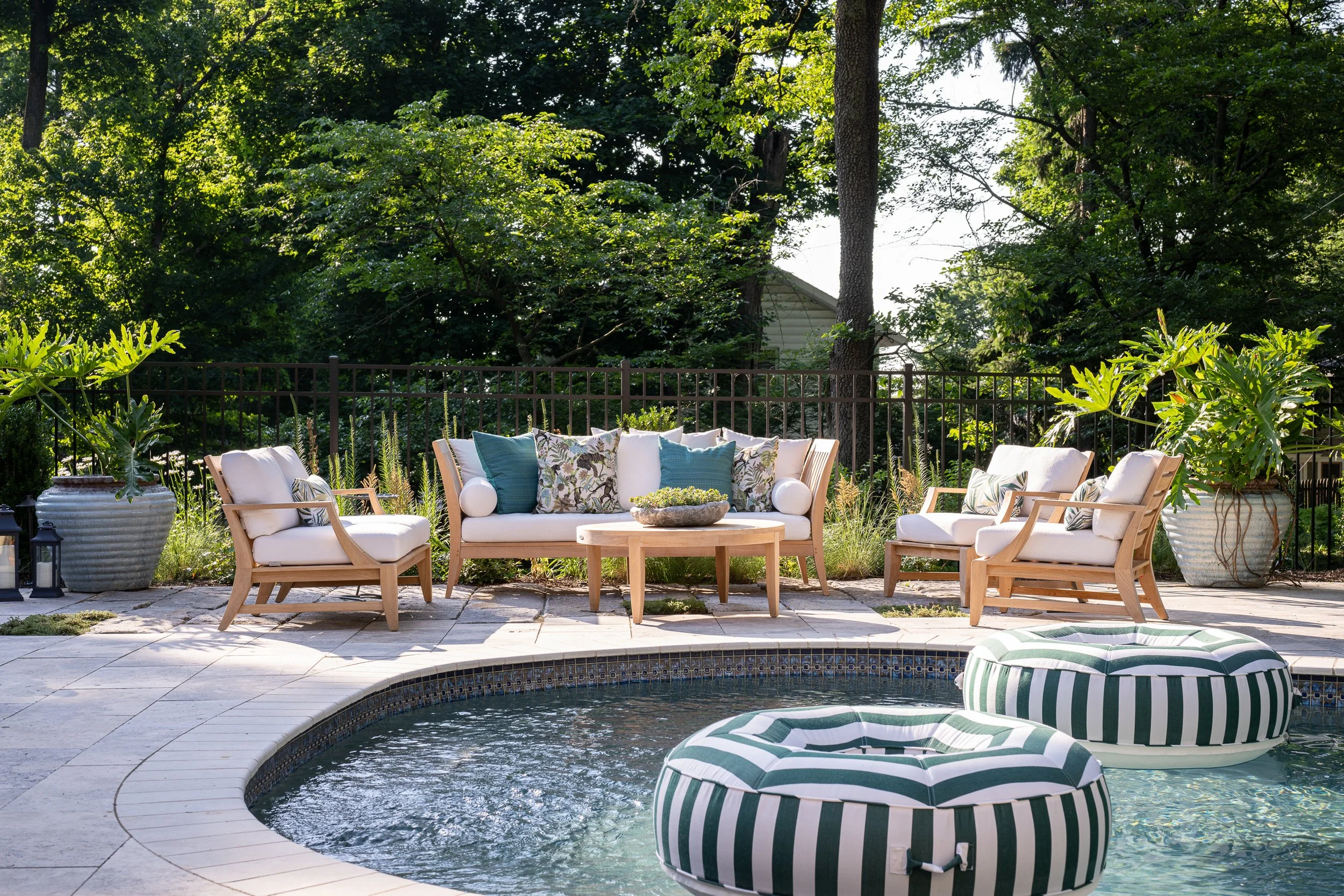 Outdoor poolside seating area with white cushioned chairs, a matching sofa with decorative pillows, and striped pool floats in the pool, surrounded by lush green trees and potted plants.
