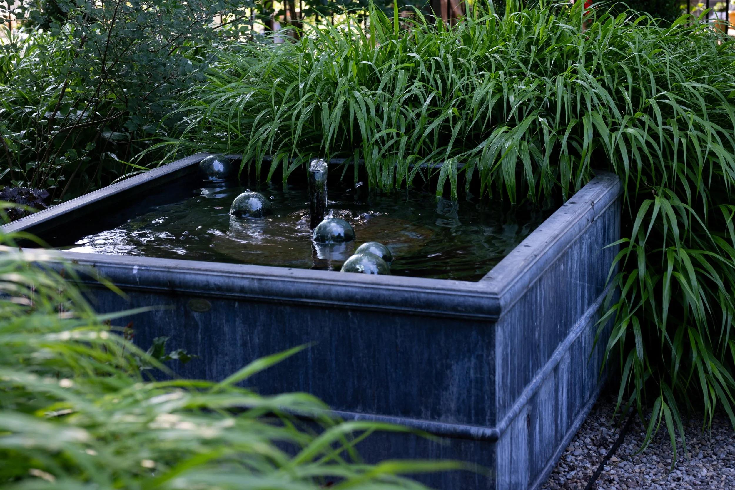 A rectangular outdoor water fountain with a central jet of water and several glass orbs, surrounded by lush green plants and tall grasses.